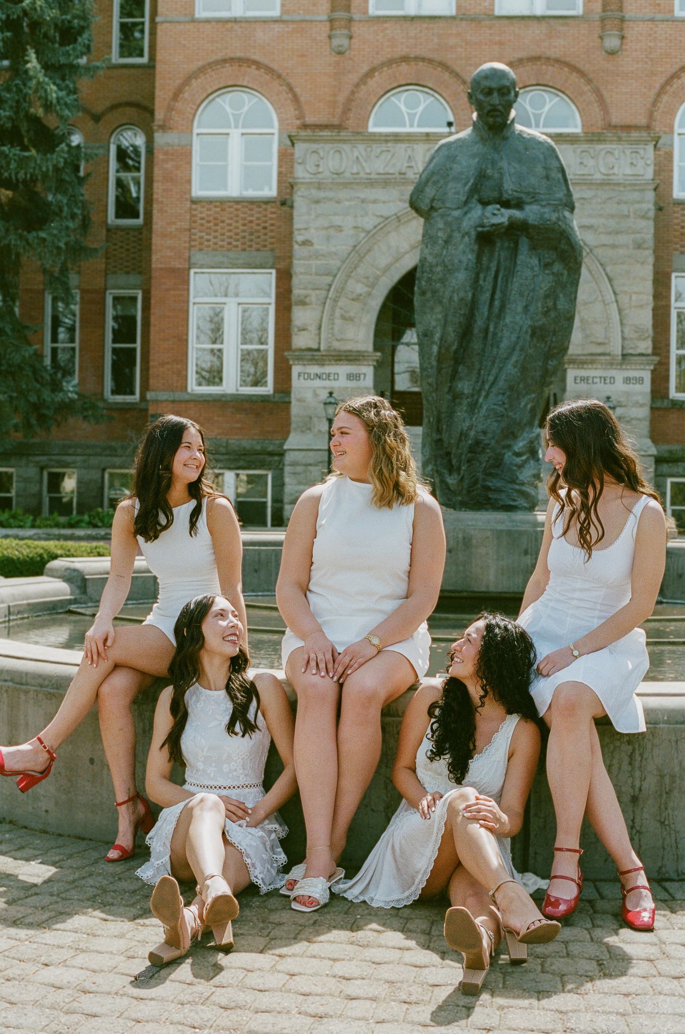 Five young women in white dresses pose together near a bronze statue outside a red brick building on a sunny day.