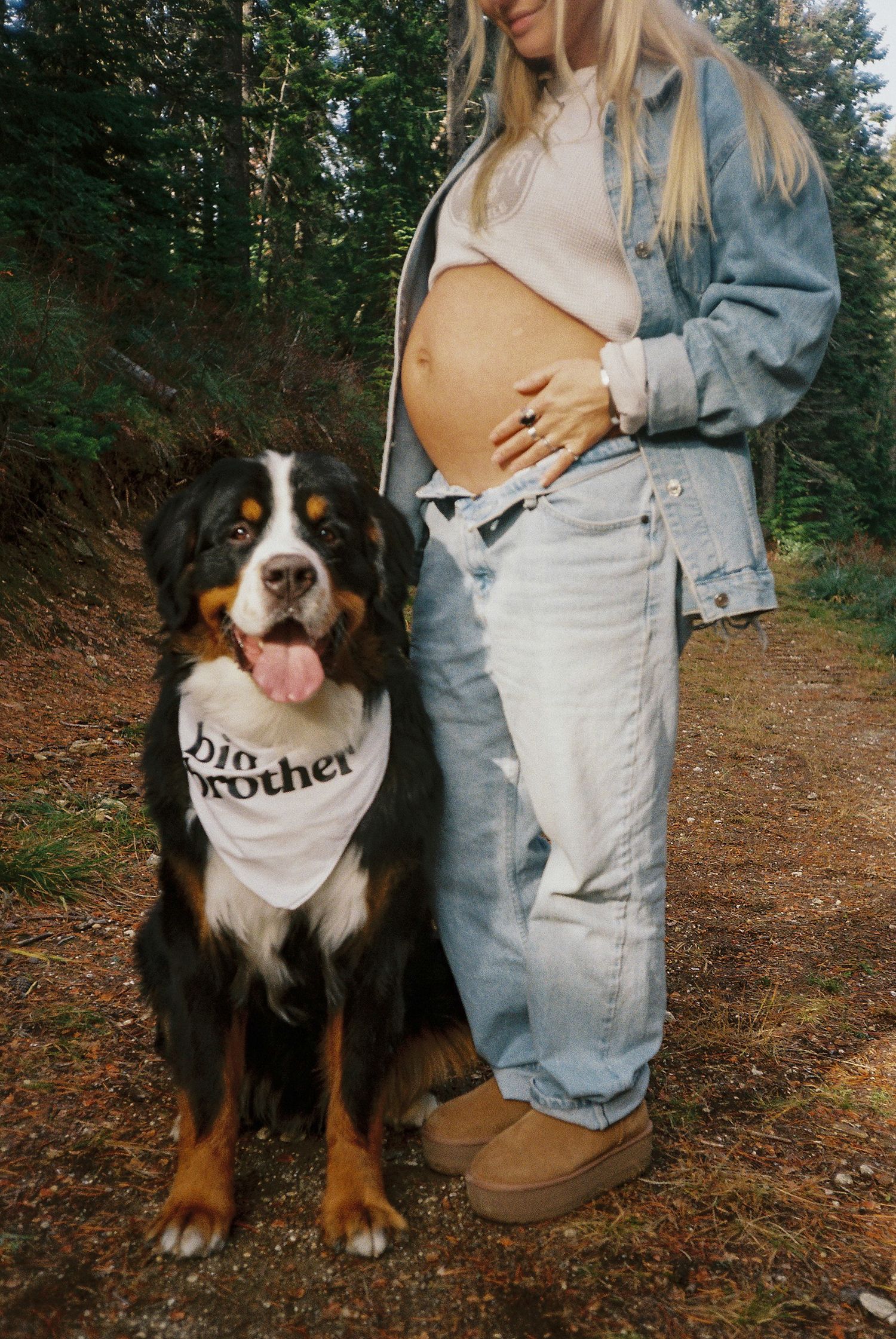 Pregnant woman in denim jacket with Bernese Mountain Dog wearing a Big Brother bandana outdoors.
