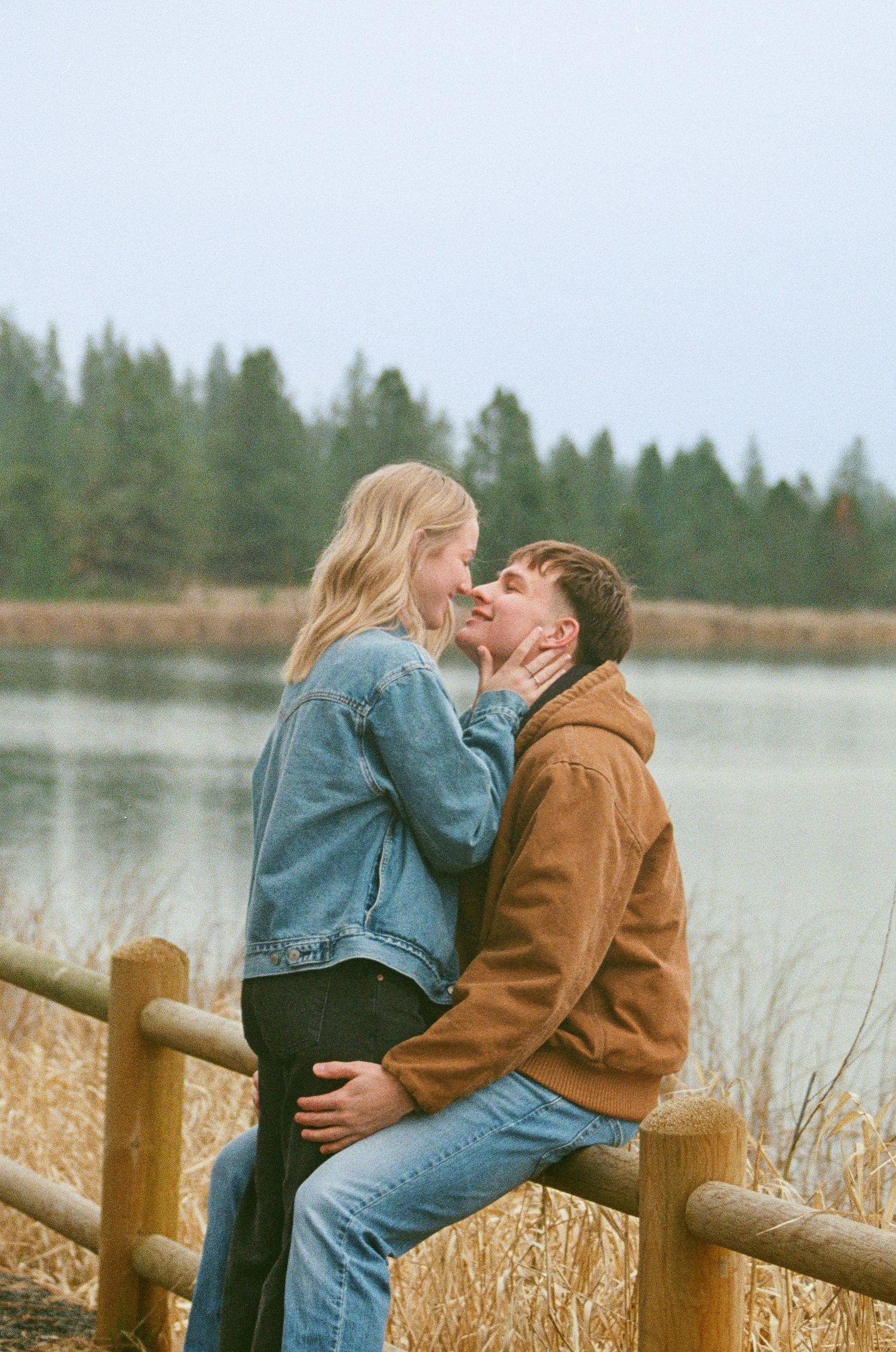 Couple sharing a romantic moment by a misty lake, woman in denim jacket sitting on wooden fence with evergreen trees behind.