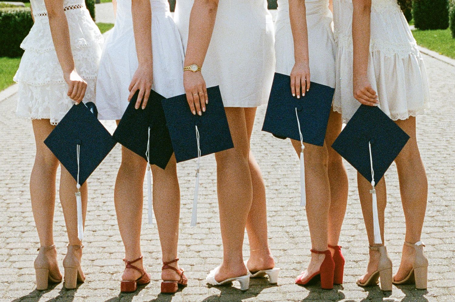 Six graduates in white dresses holding navy blue mortarboard caps, wearing heels, posing together outdoors.