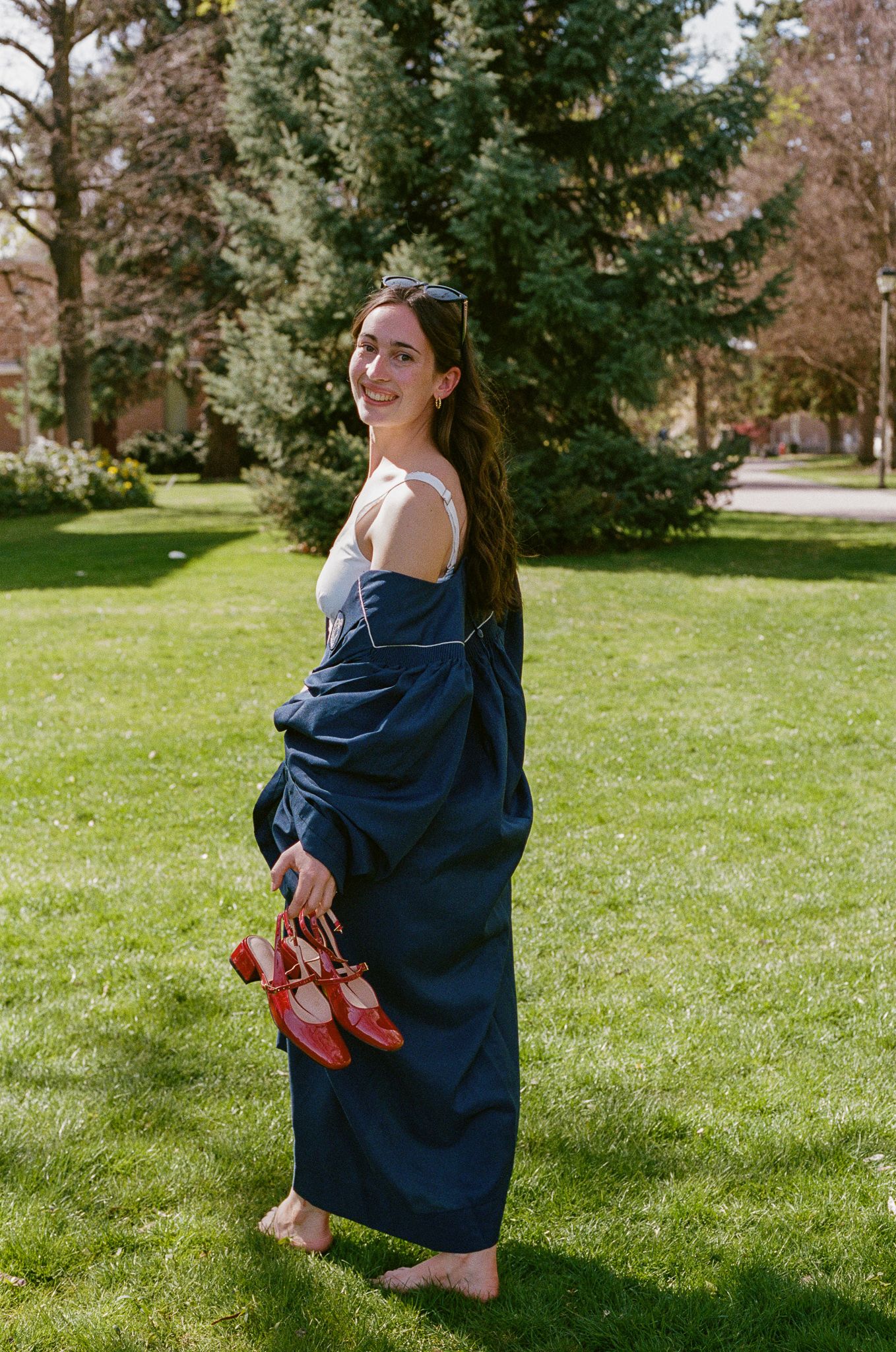 Smiling female graduate in navy robe stands barefoot on grass, holding red heels, posing outdoors.