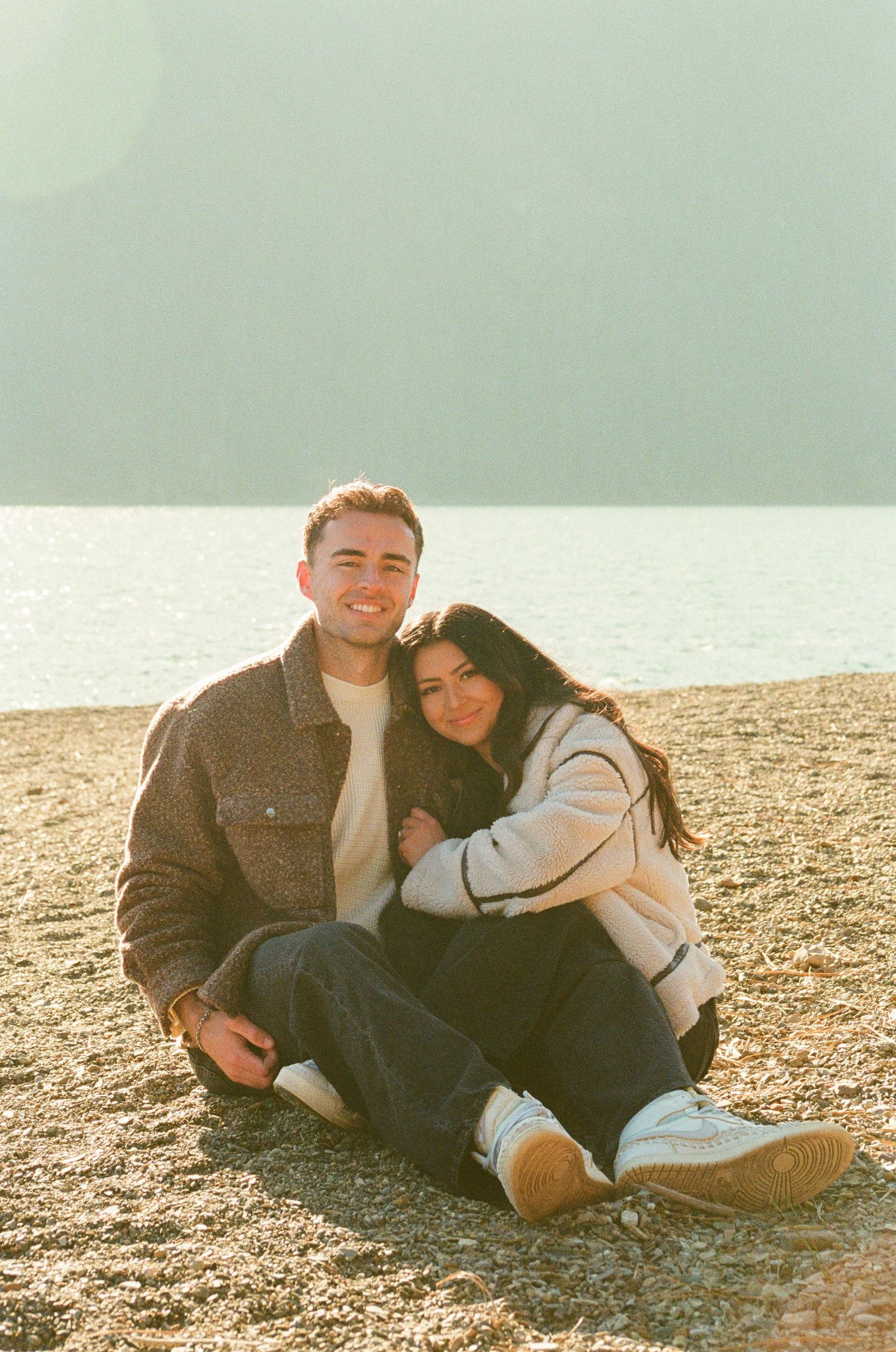 A smiling couple sits together on a rocky beach shore with a scenic mountain lake backdrop in warm golden light.
