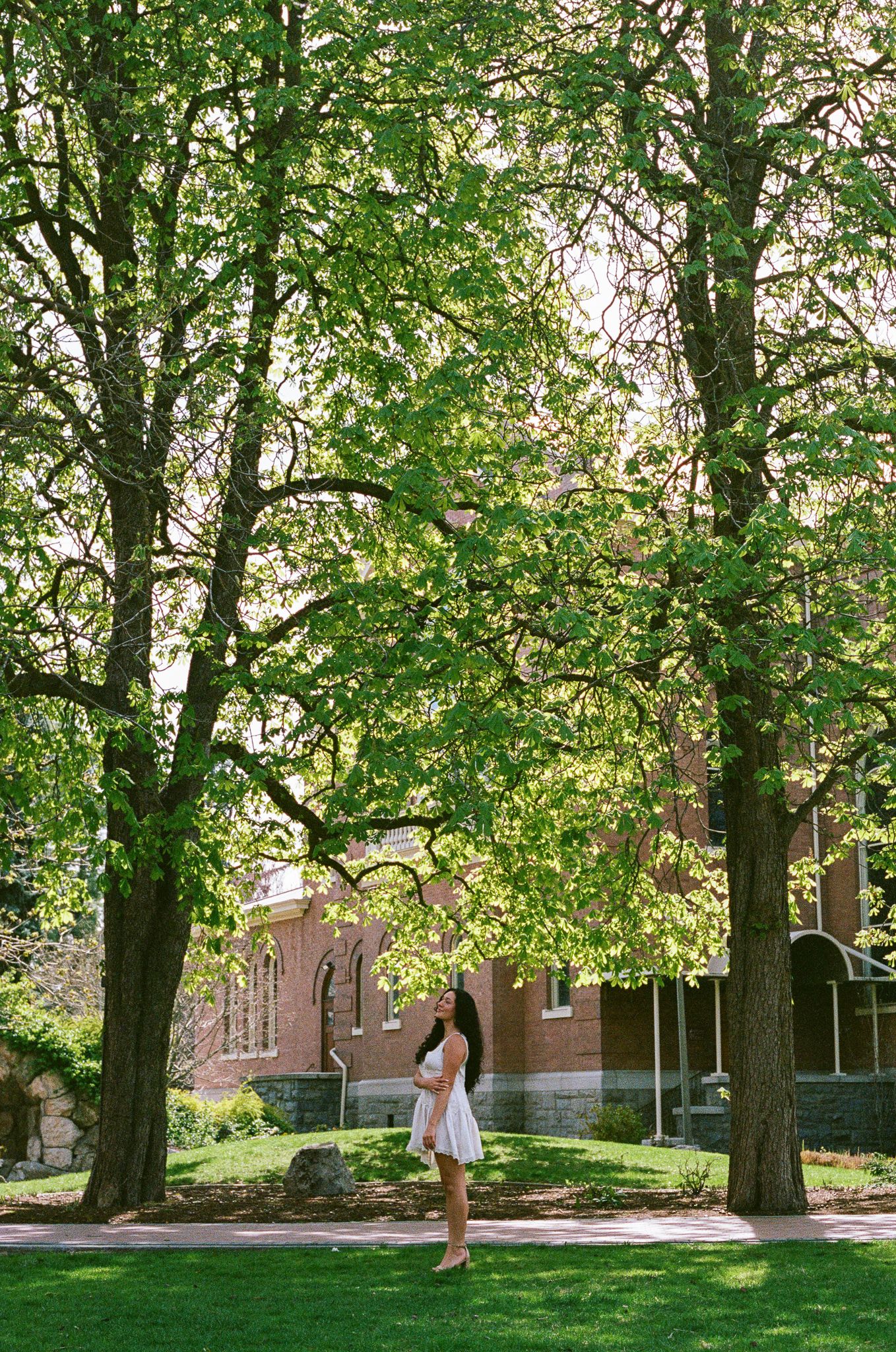 Woman in white dress standing beneath tall green trees on a sunny day near a red brick building on a college campus.