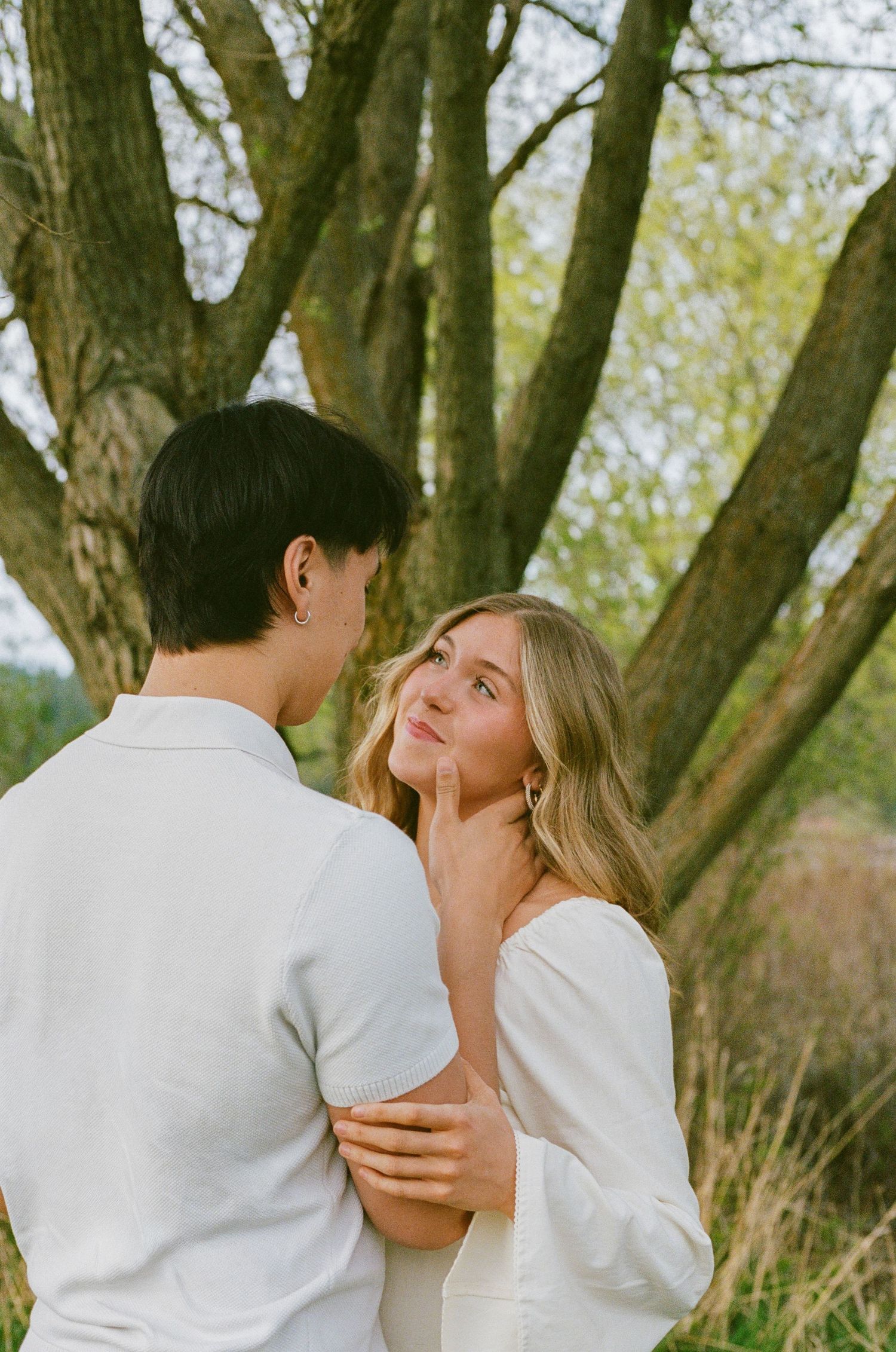 Couple in white outfits share a tender moment under a large tree, woman gazing up lovingly at her partner.