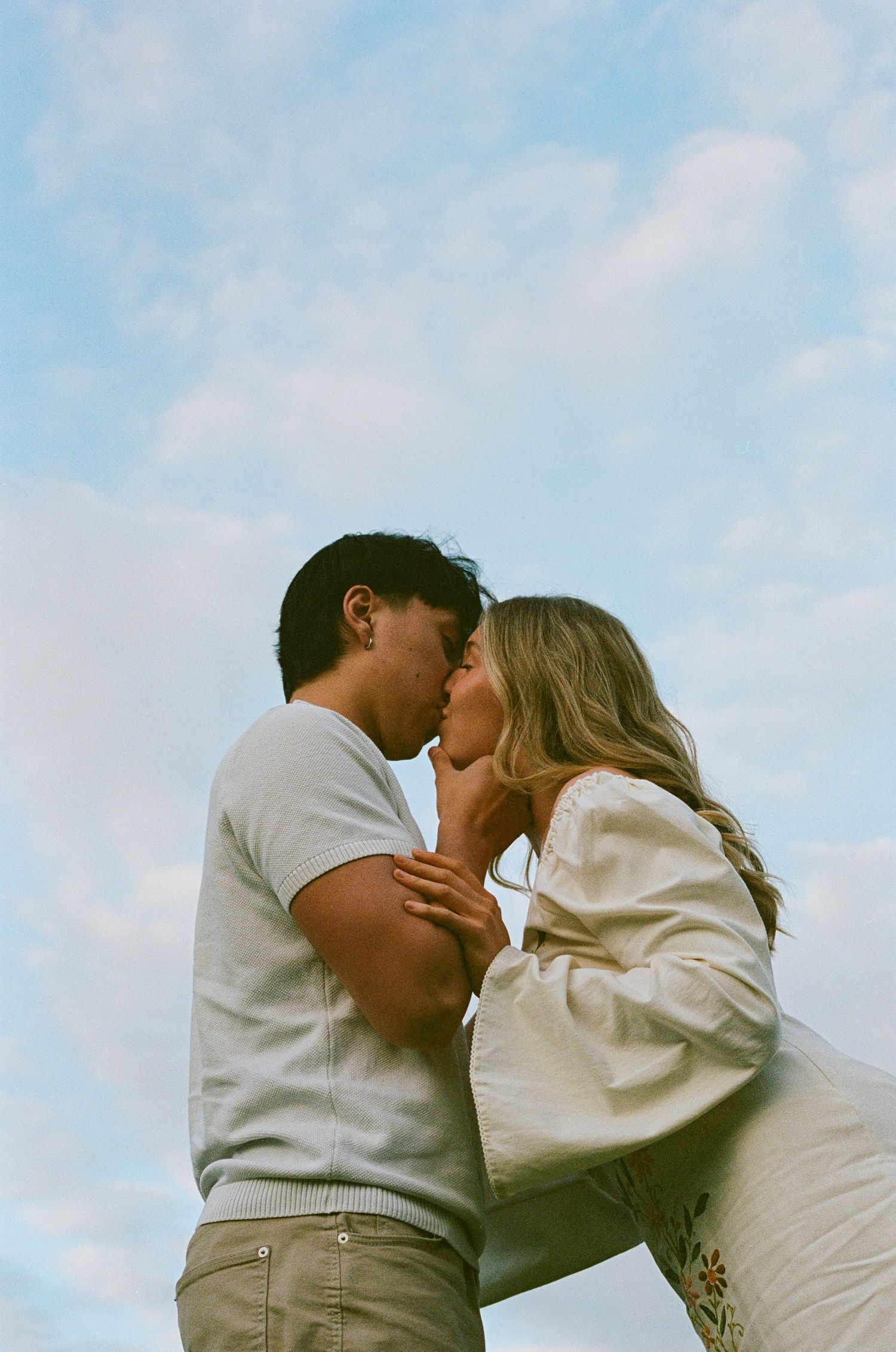 A couple shares a romantic kiss outdoors against a blue cloudy sky, dressed in casual white and beige outfits.