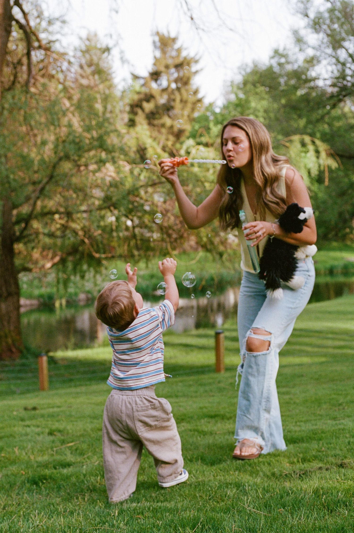A young woman blows bubbles for a toddler outdoors, holding a small black cat in a lush green backyard.