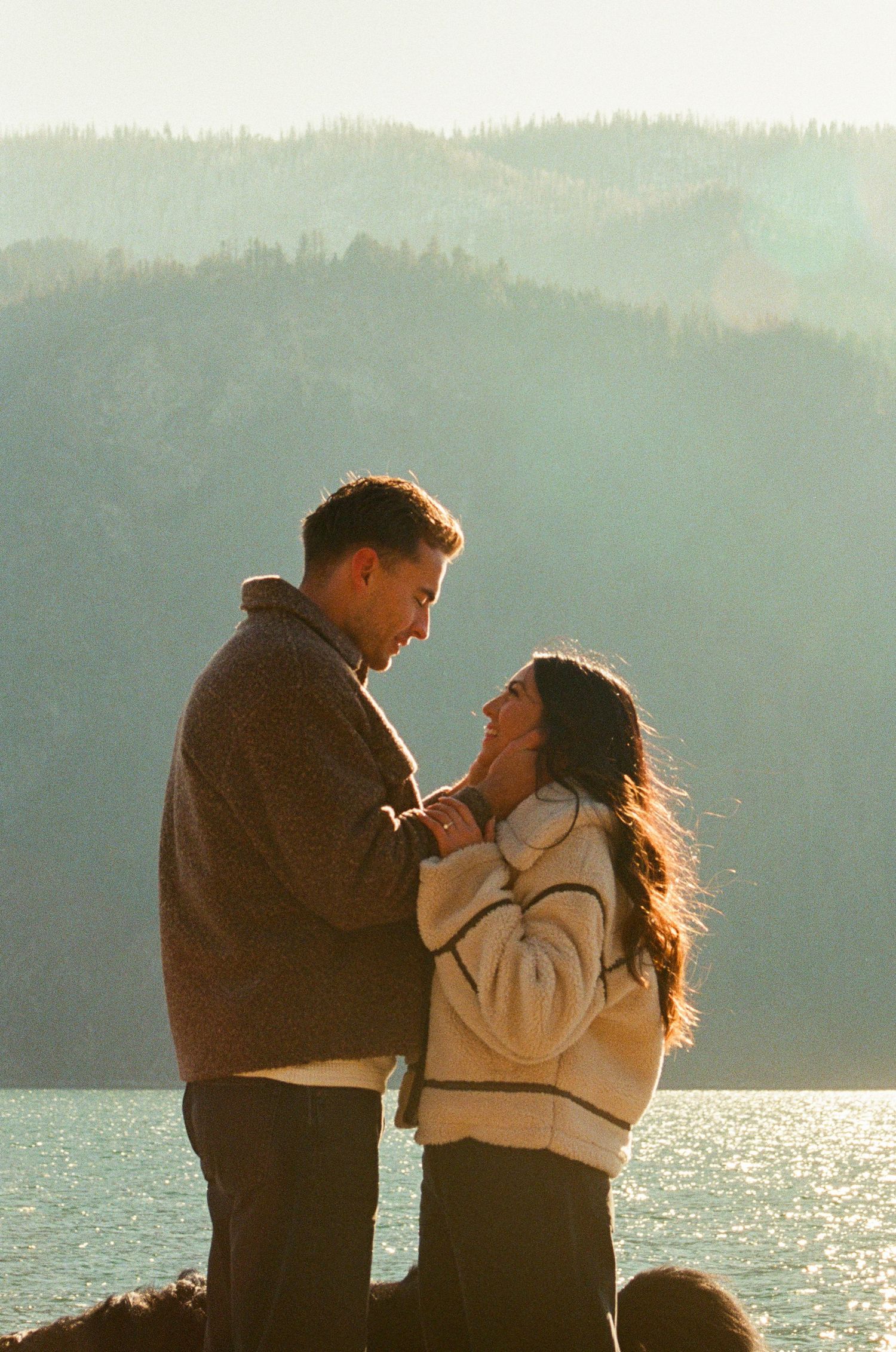 Couple embracing by a misty mountain lake at golden hour, wearing cozy sweaters in a romantic outdoor setting.