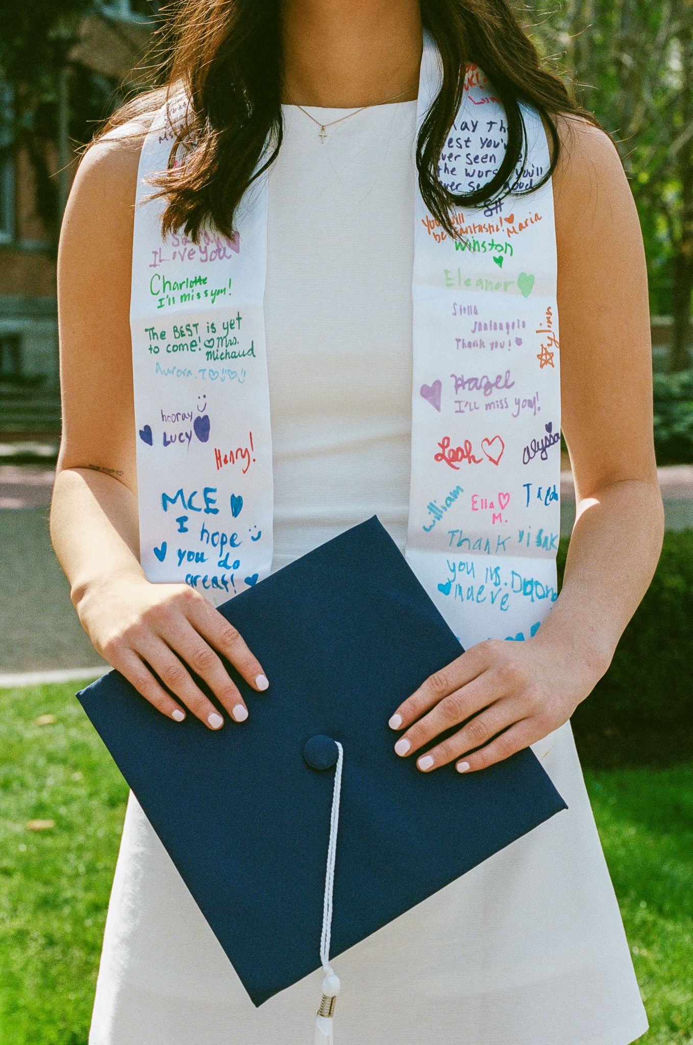 Graduate in white dress holds navy cap, wearing a signed white graduation stole with colorful messages from friends.