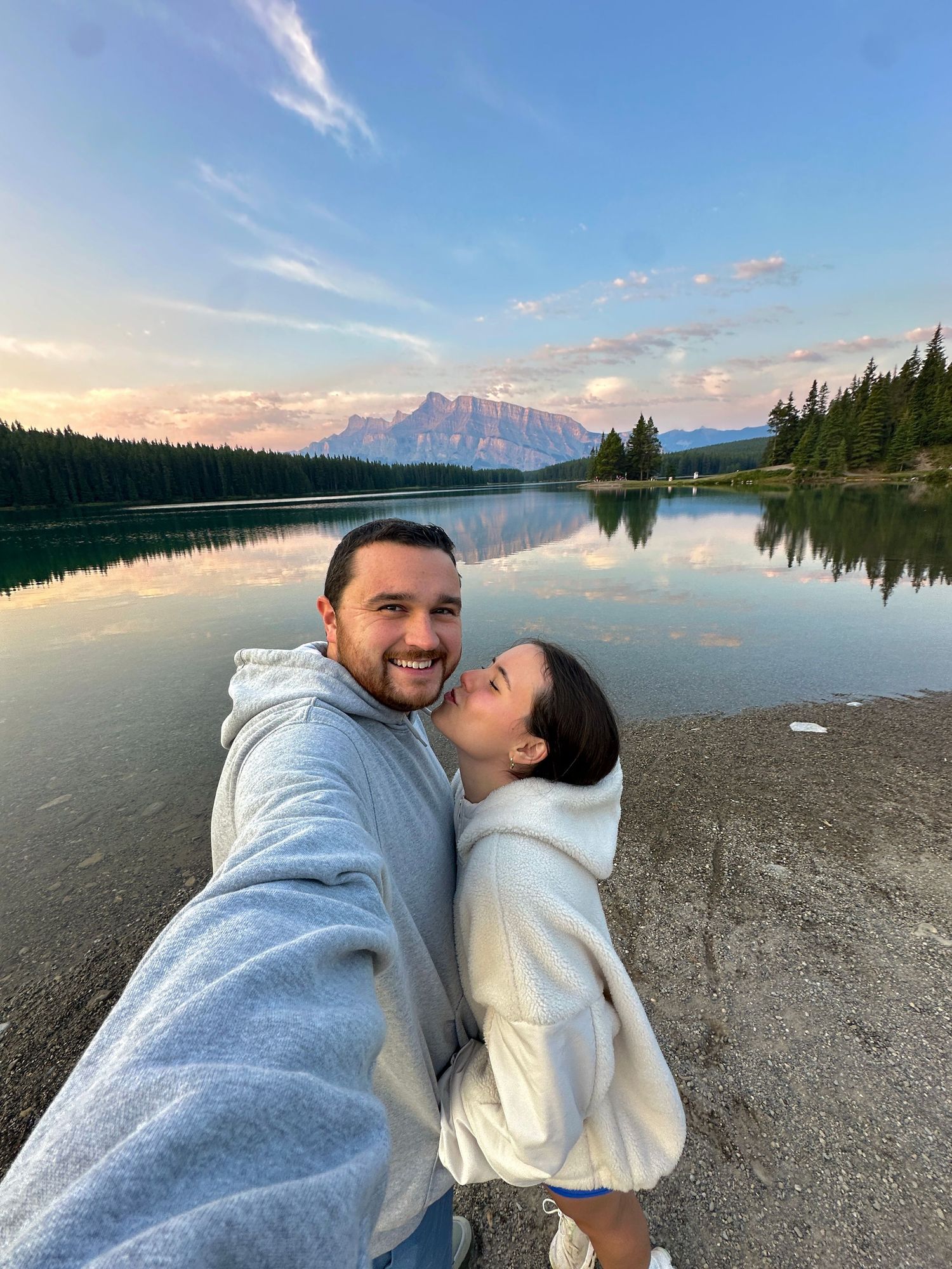 Couple taking selfies on a rocky lakeshore with mountain views at sunset.