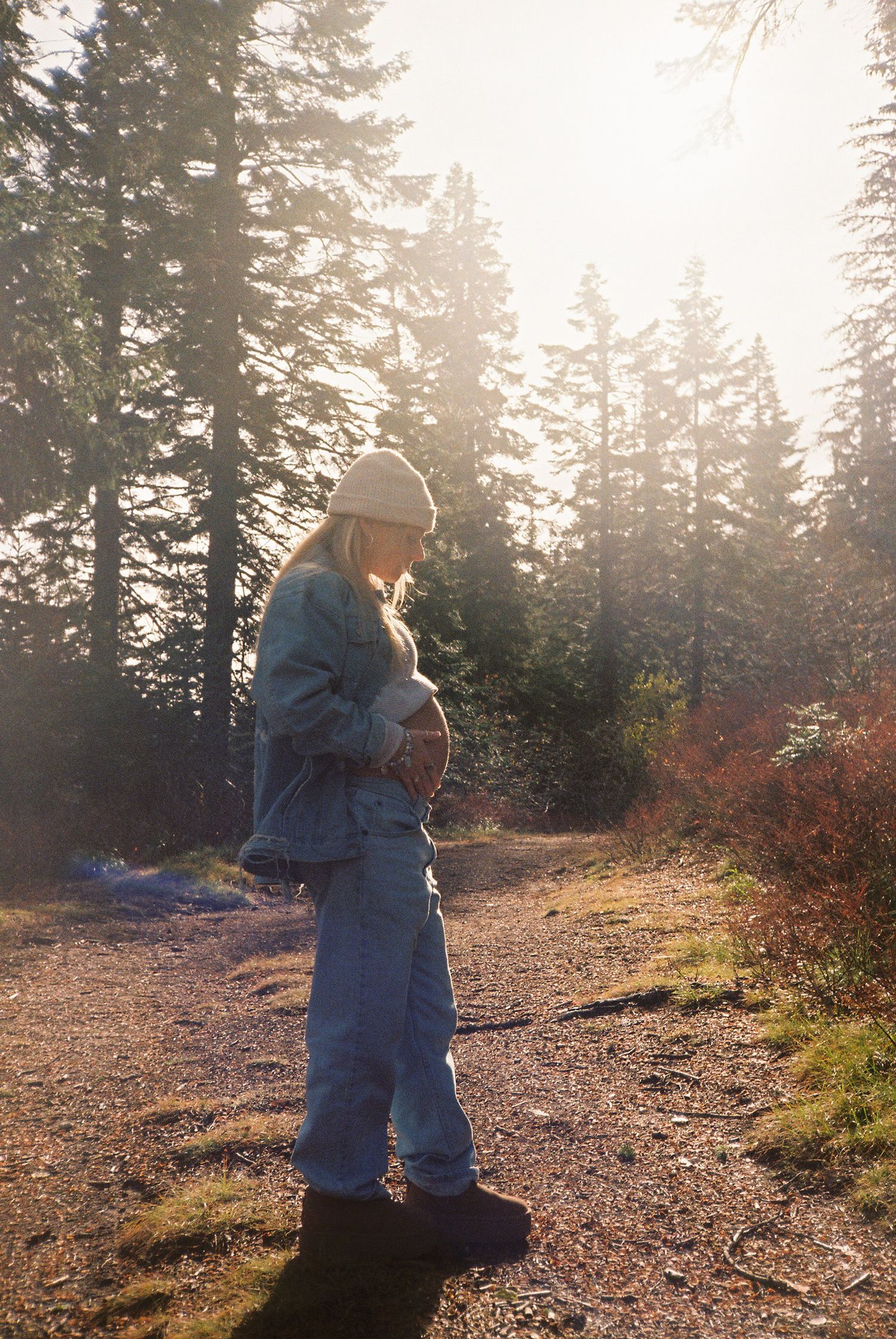 Pregnant woman in denim jacket and beanie cradles baby bump on sunlit forest trail surrounded by tall pine trees.
