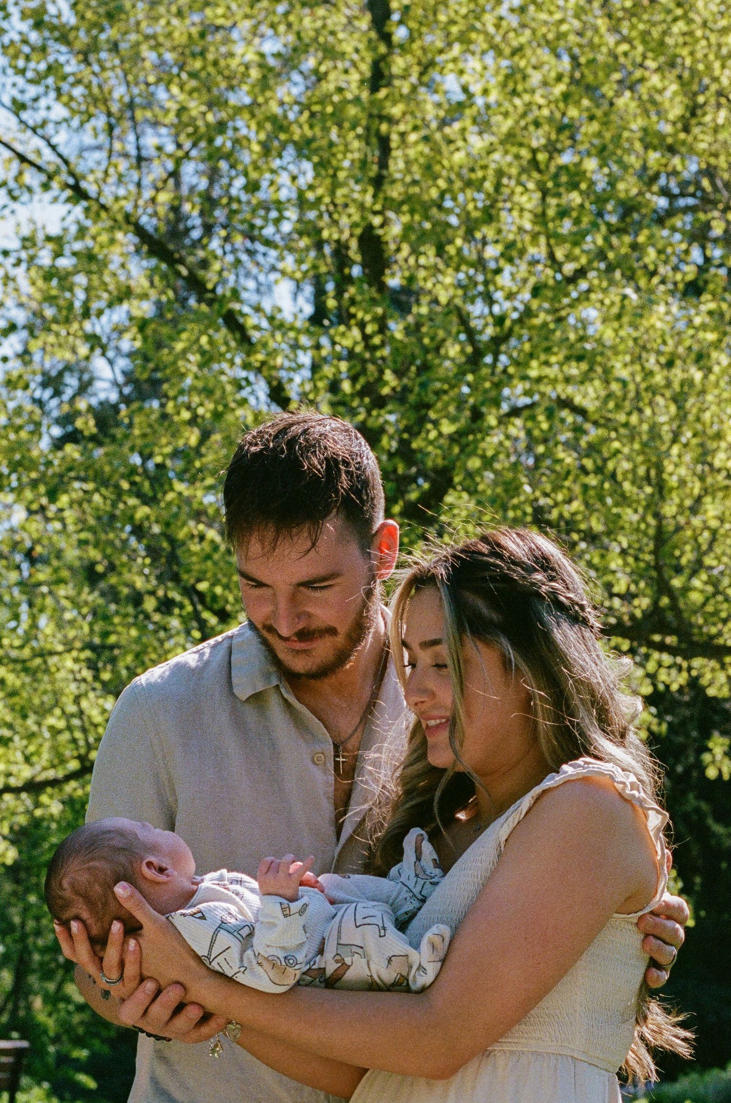 A couple tenderly holds their newborn baby outdoors surrounded by lush green trees on a sunny day.