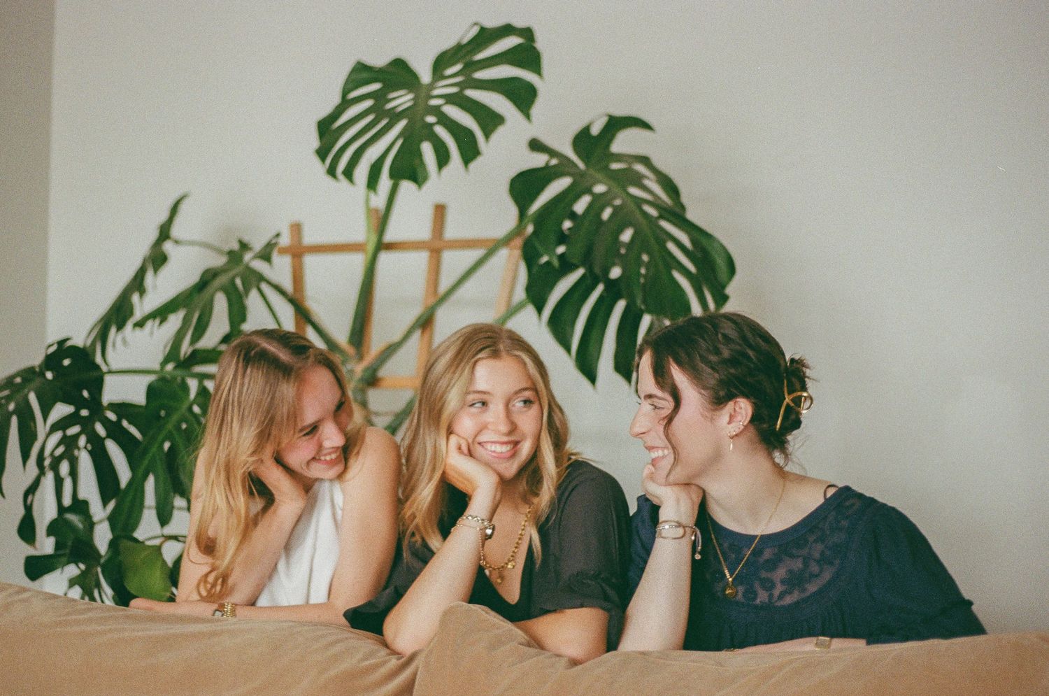 Three young women smiling and laughing together behind a couch with a monstera plant in the background.