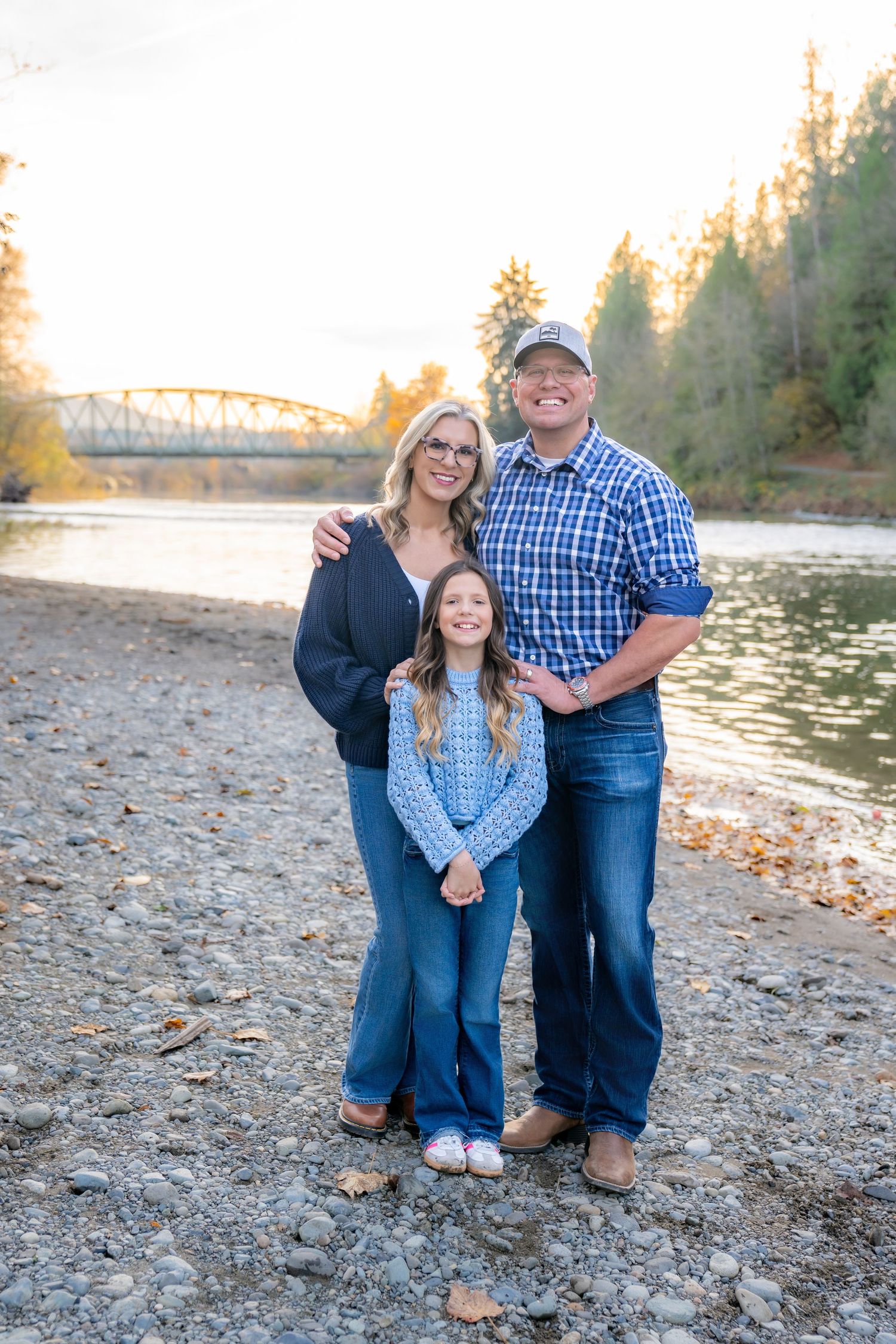 Family portrait on rocky riverbank with bridge and autumn trees in background.