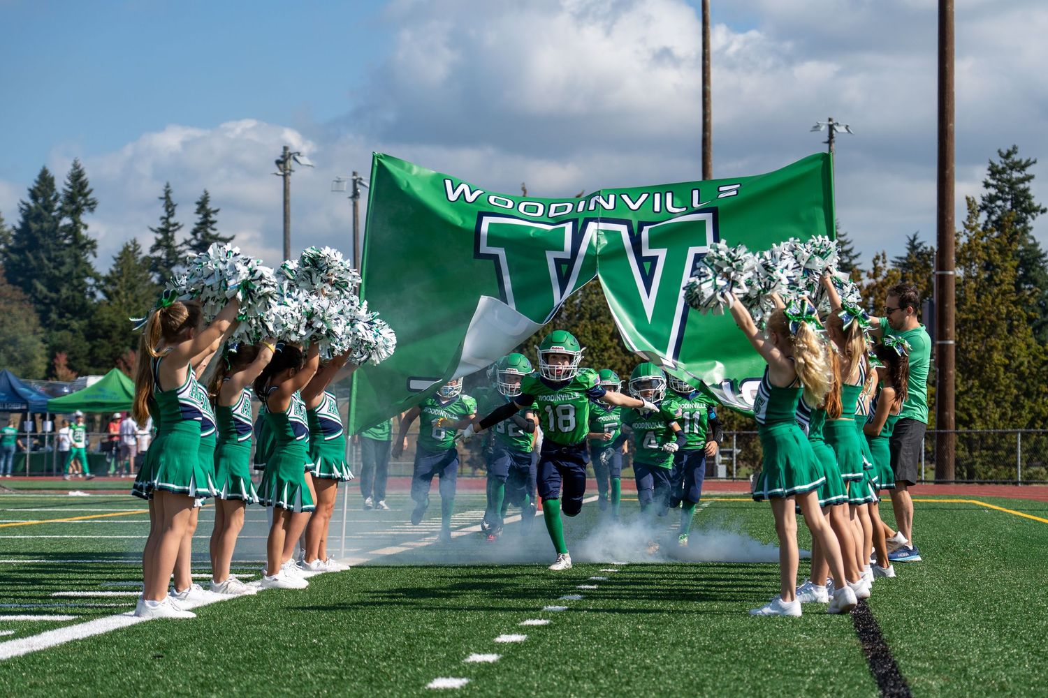 Cheerleaders form tunnel entrance for football team through green banner on field.