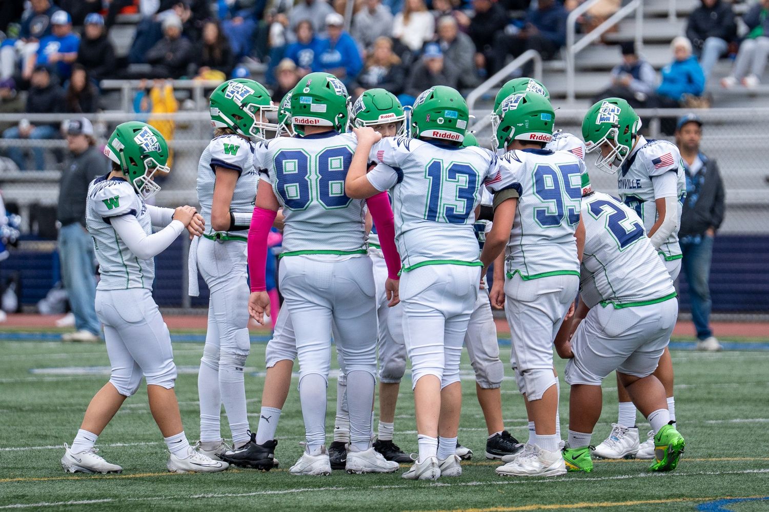 Football team huddles on field wearing green uniforms with pink breast cancer awareness accessories.