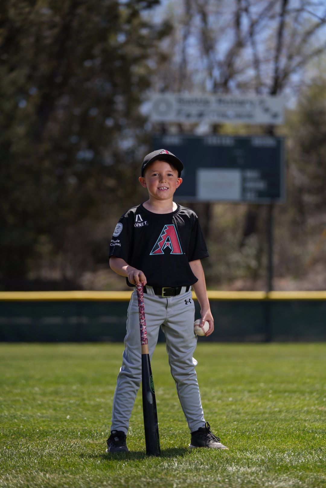 Young baseball player stands with bat on field wearing Arizona Diamondbacks uniform and cap during sunny day.