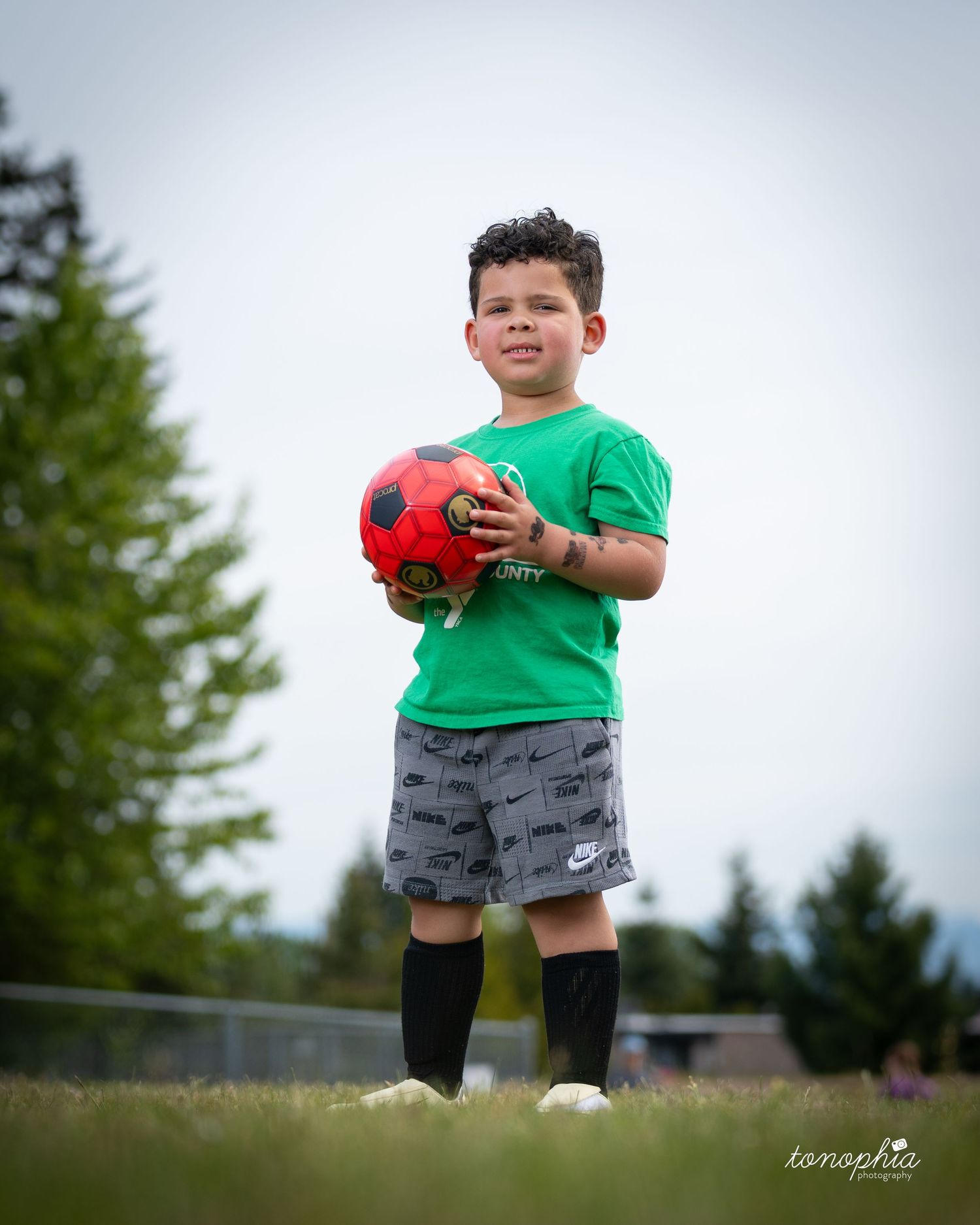 A child in a green shirt and gray shorts holds a red ball while standing in a grassy field.