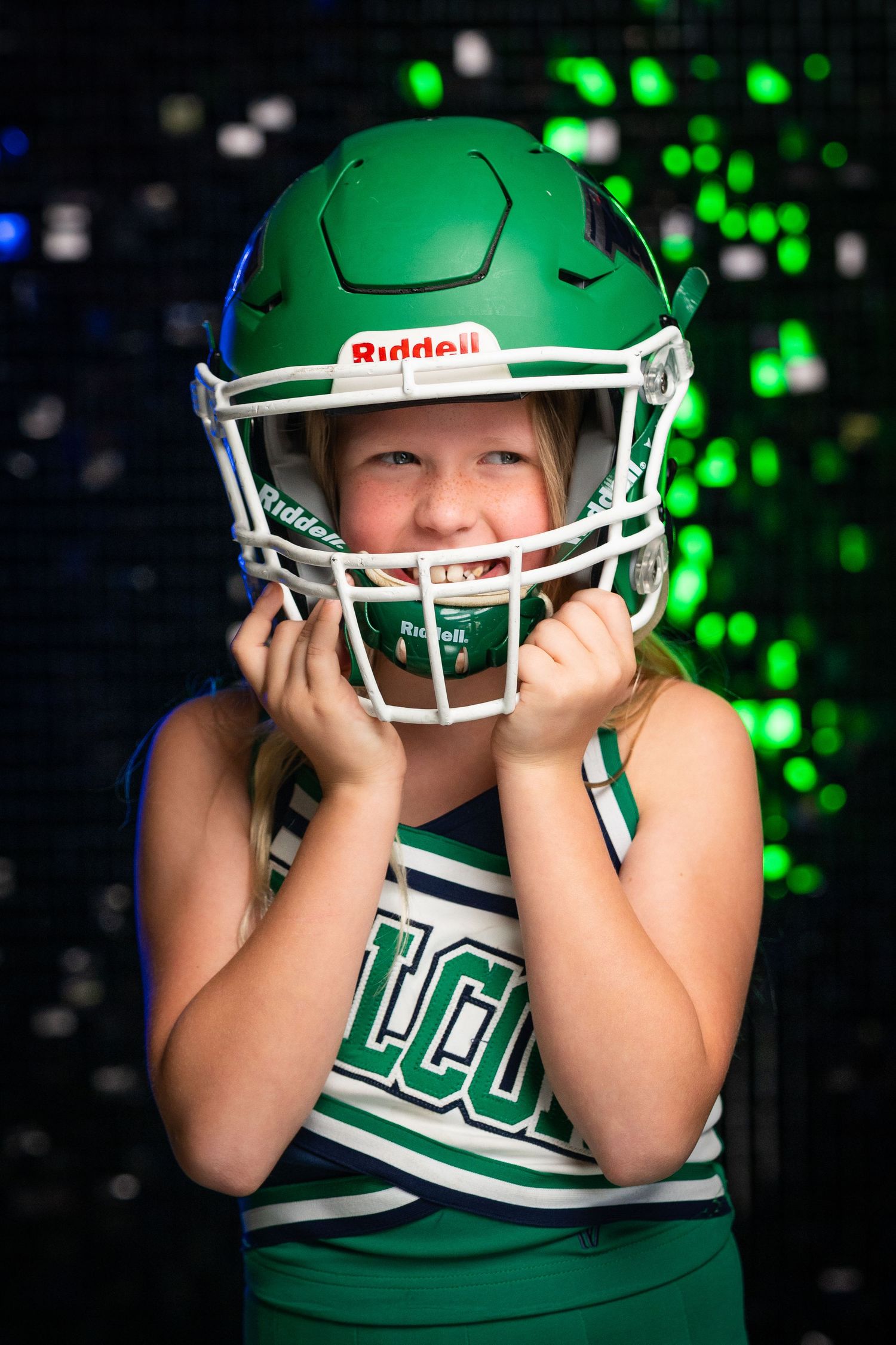 Youth cheerleader wearing green Riddell helmet poses against sparkly background.