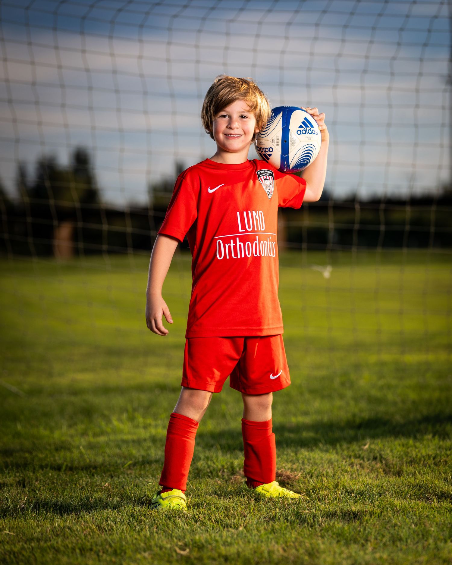 Young soccer player in red uniform stands on green grass field with goal net in background during sunny day.