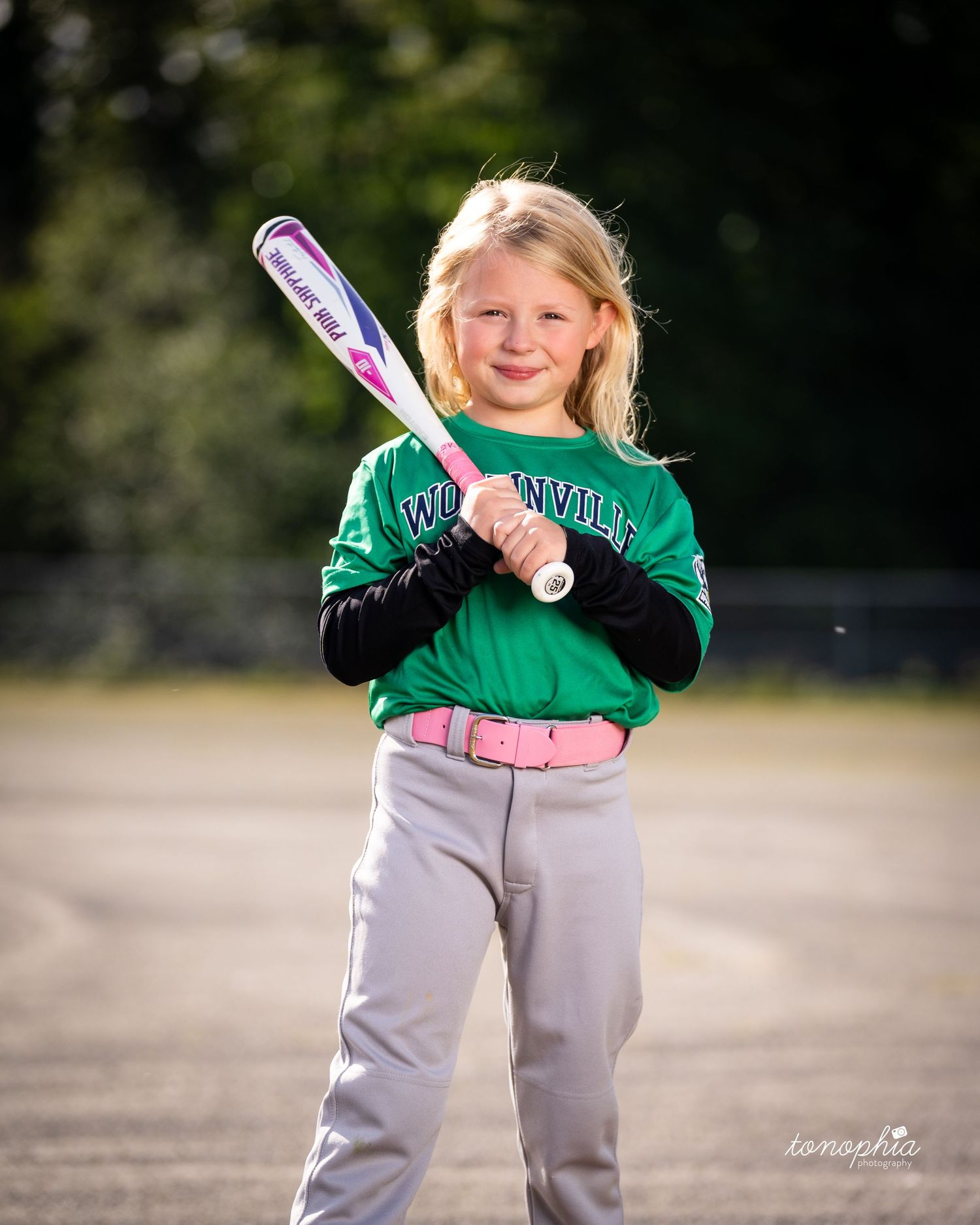 Young softball player in a green and white uniform holds a pink bat while smiling outdoors on a sunny day.