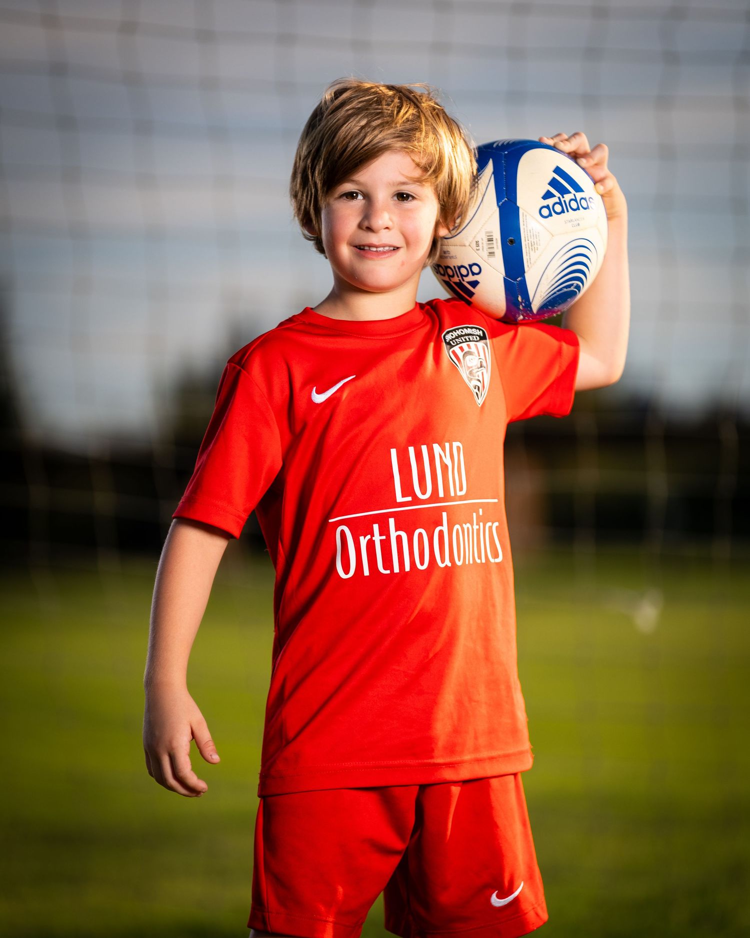 Young soccer player in red uniform poses with a ball on a sunny grass field during practice.