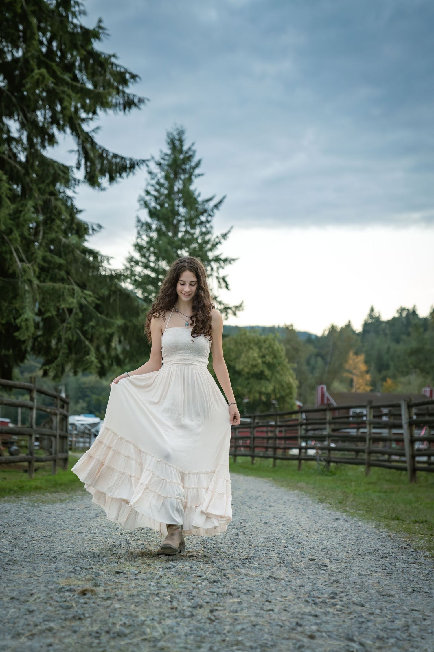 Person in a flowing white dress twirls on a gravel path surrounded by evergreen trees.