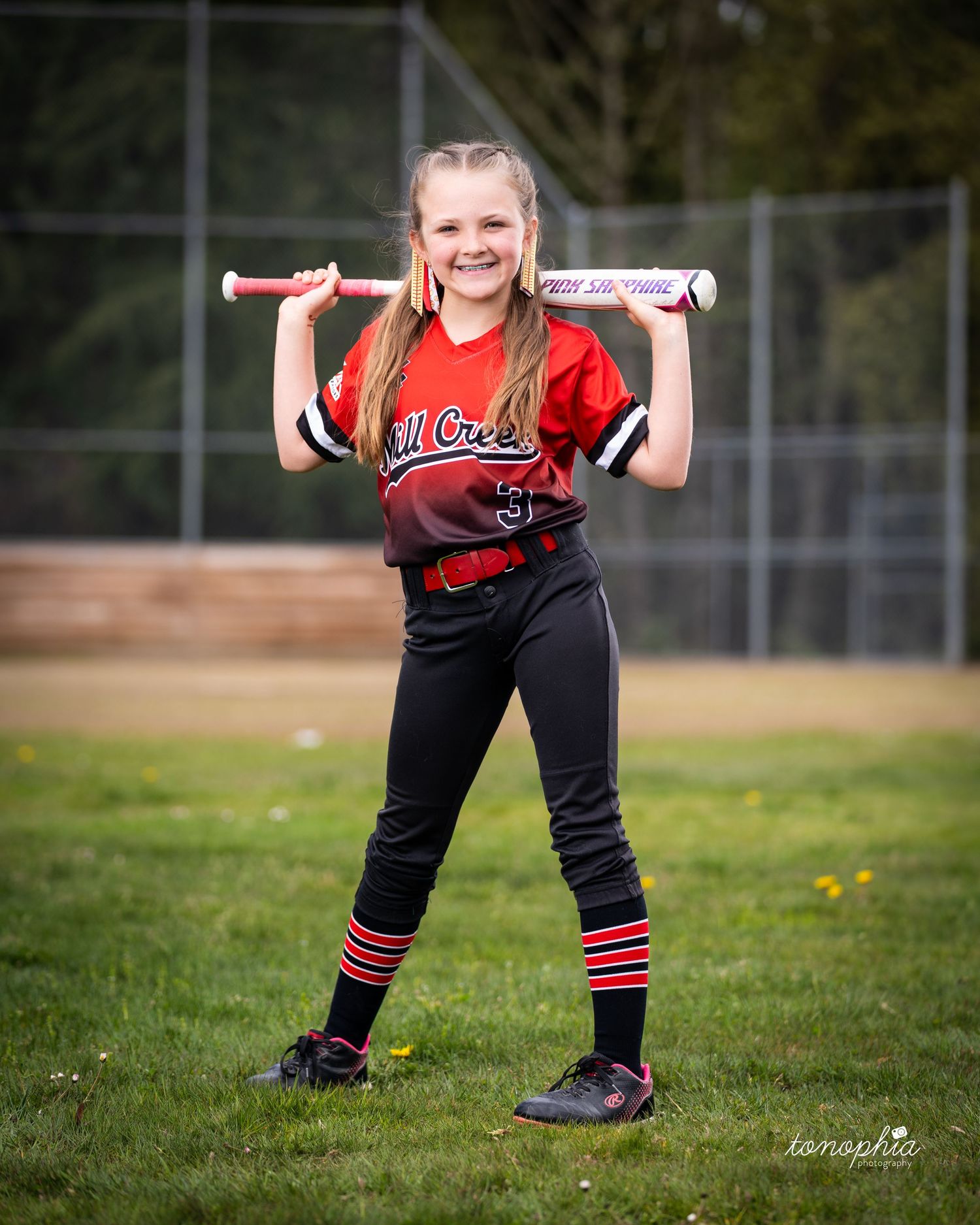 A softball player in red and black uniform crouches in a batting stance on a grassy field during practice.