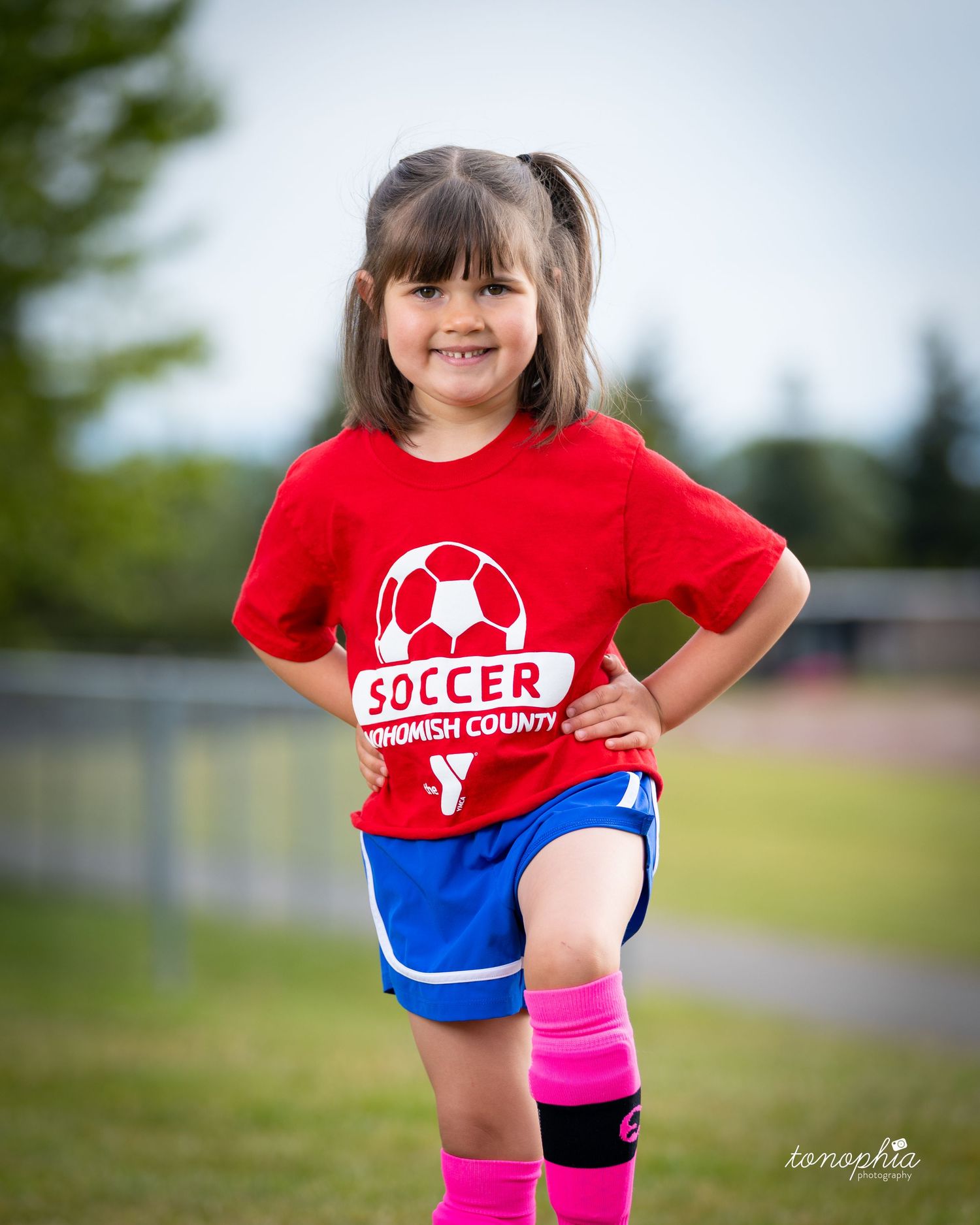 Young athlete in red soccer shirt and blue shorts poses playfully outdoors on sunny field.