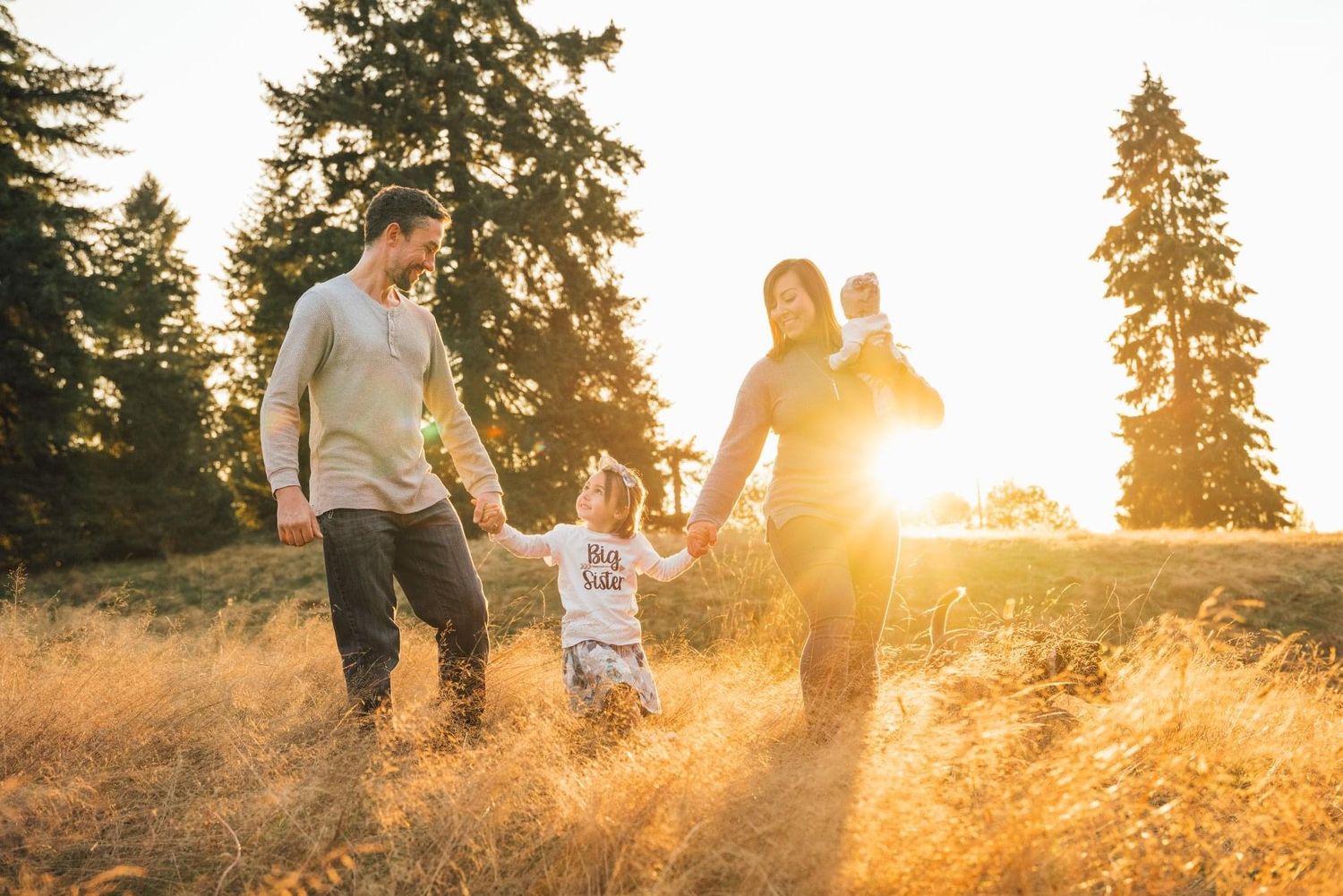 Silhouettes of people walking through golden grass field at sunset with tall pine trees.