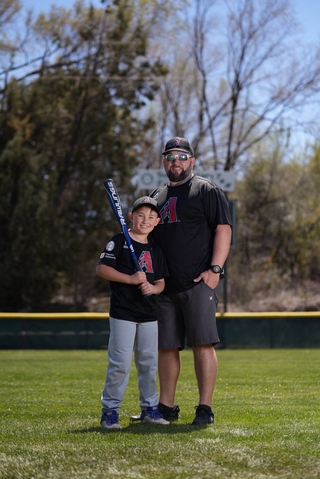 Baseball coach and young player pose together on sunny field with gloves and equipment.