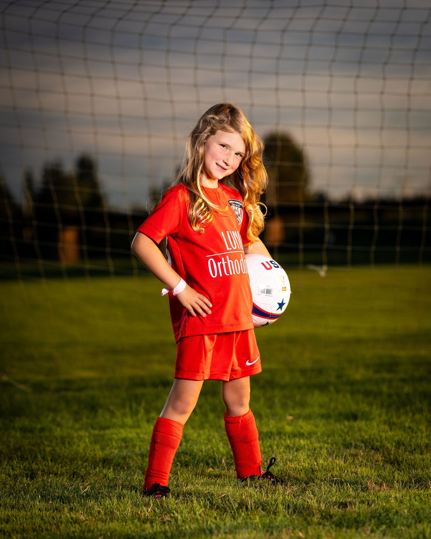 Young soccer player in red uniform practicing with a ball on a grassy field at sunset.