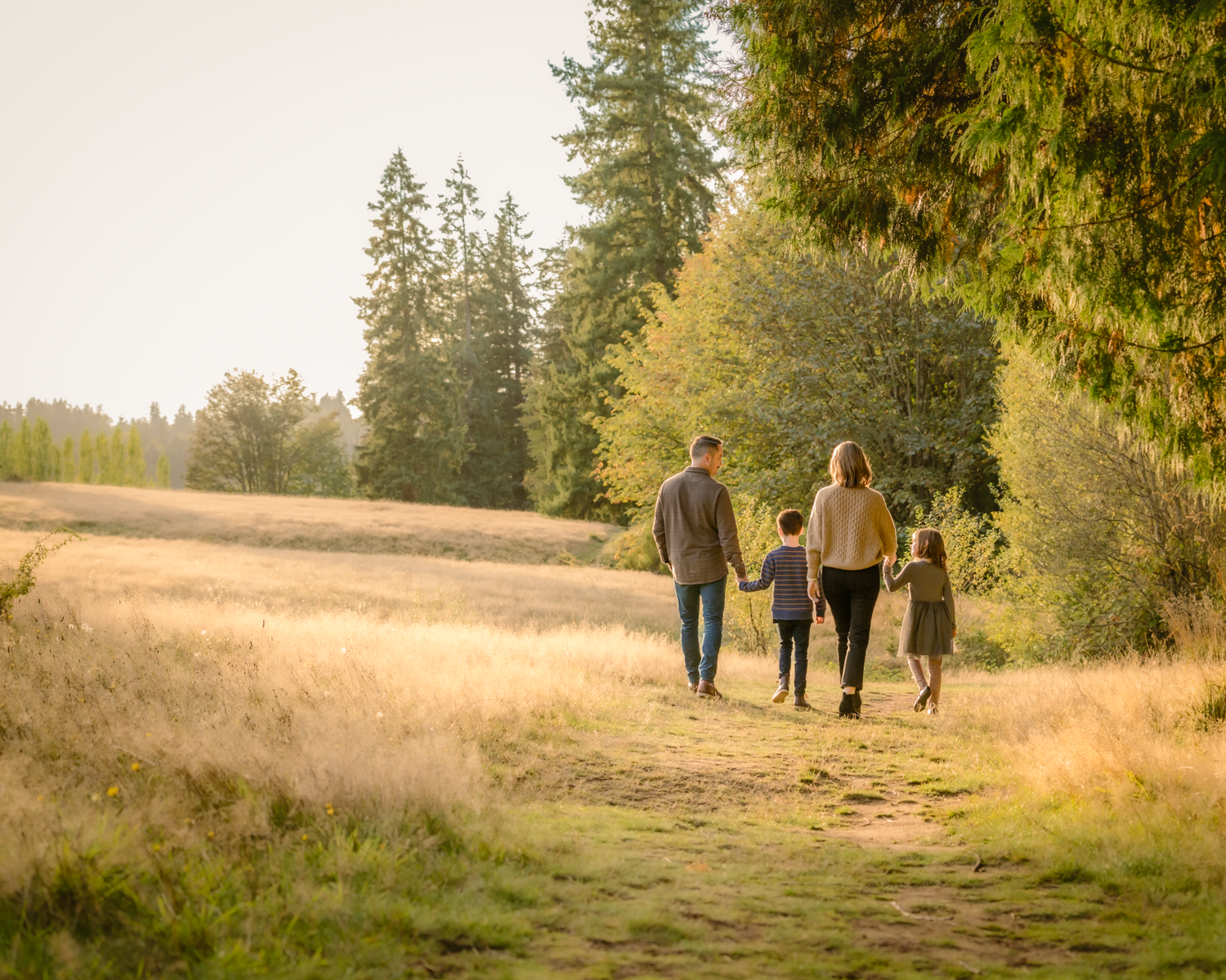 Family walking together on nature trail through golden autumn field with evergreen forest backdrop.