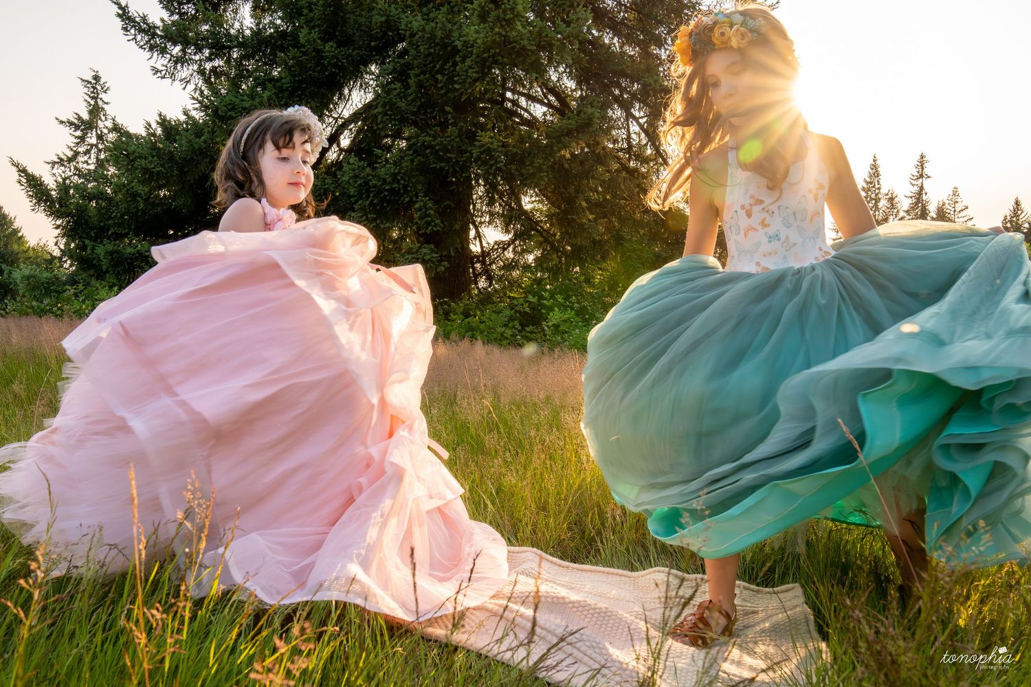 Elegant ballgowns swirl through a sunlit field with evergreen trees in the background.
