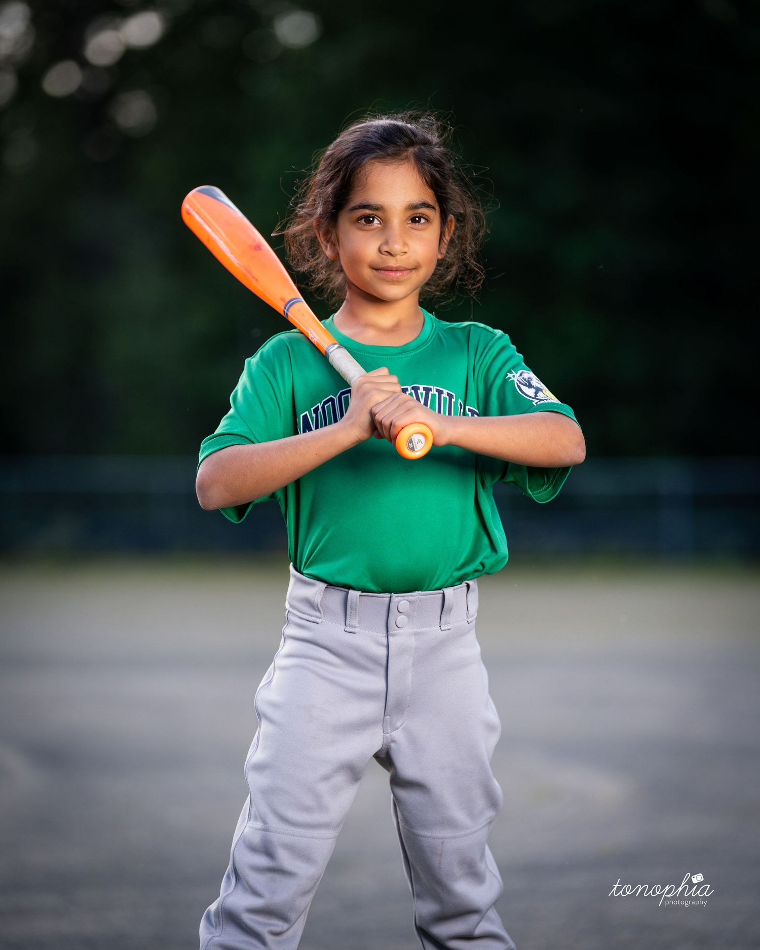 Young baseball player in green jersey and gray pants poses with orange bat on field at dusk.