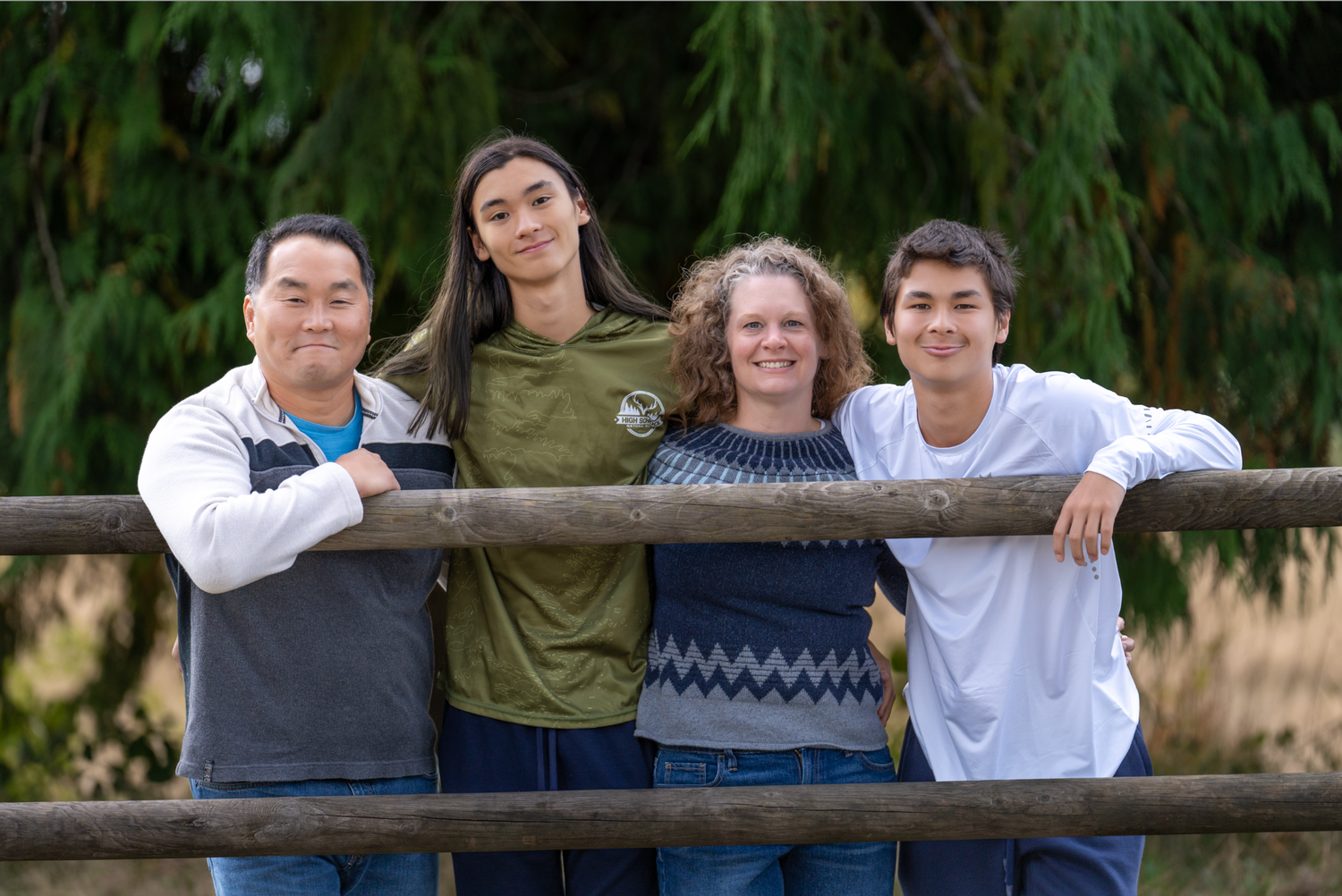 Family group poses together on a wooden fence at sunset with evergreen trees in the background.