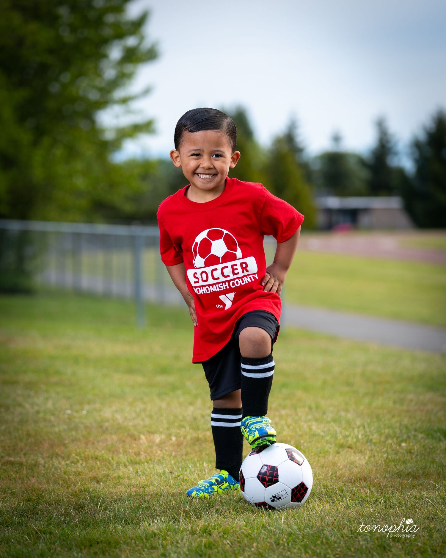 A young soccer player in a red jersey and black shorts practices dribbling with a soccer ball on a grassy field.