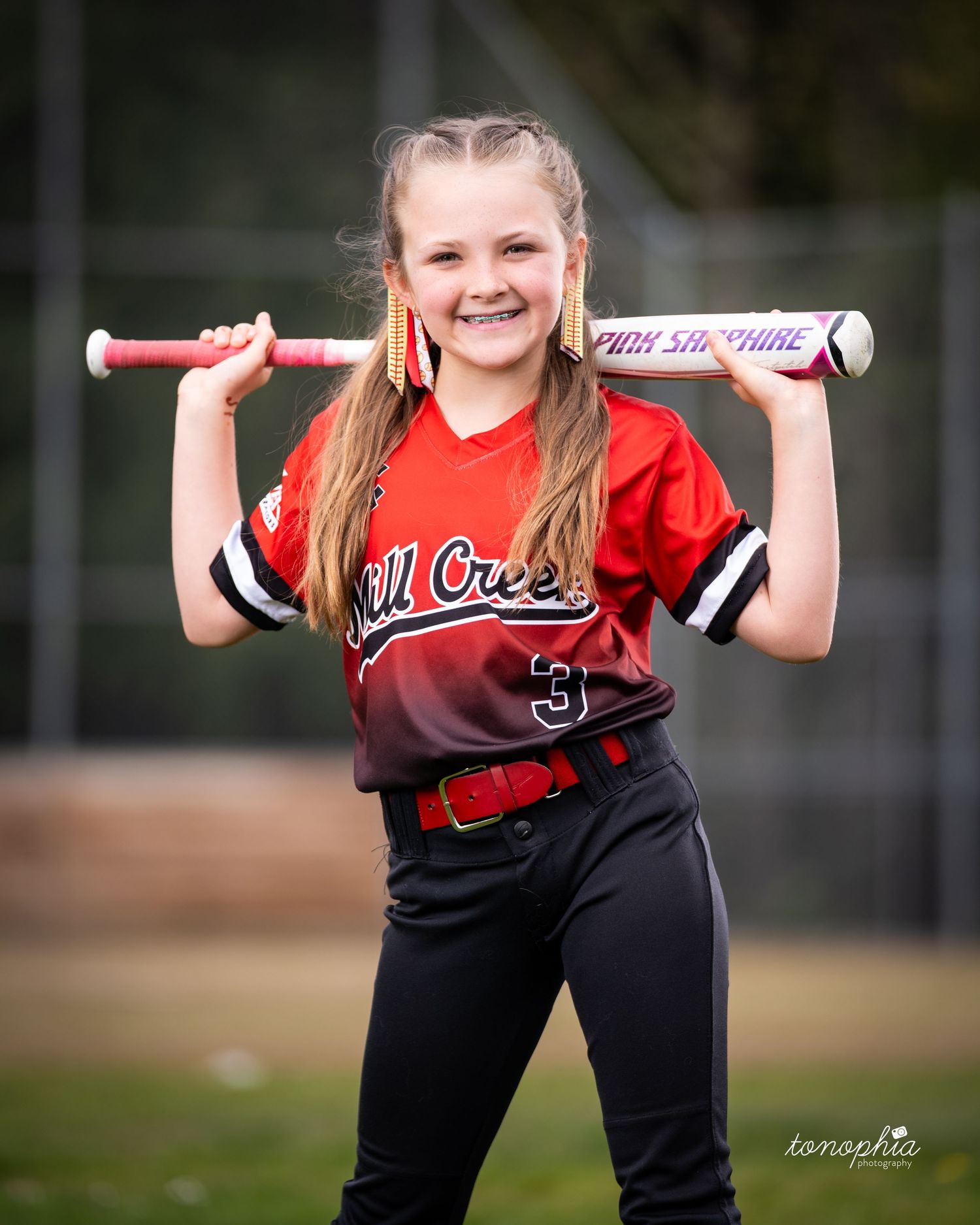 Youth softball player in red and black uniform poses with bat resting on shoulders during team photo session.