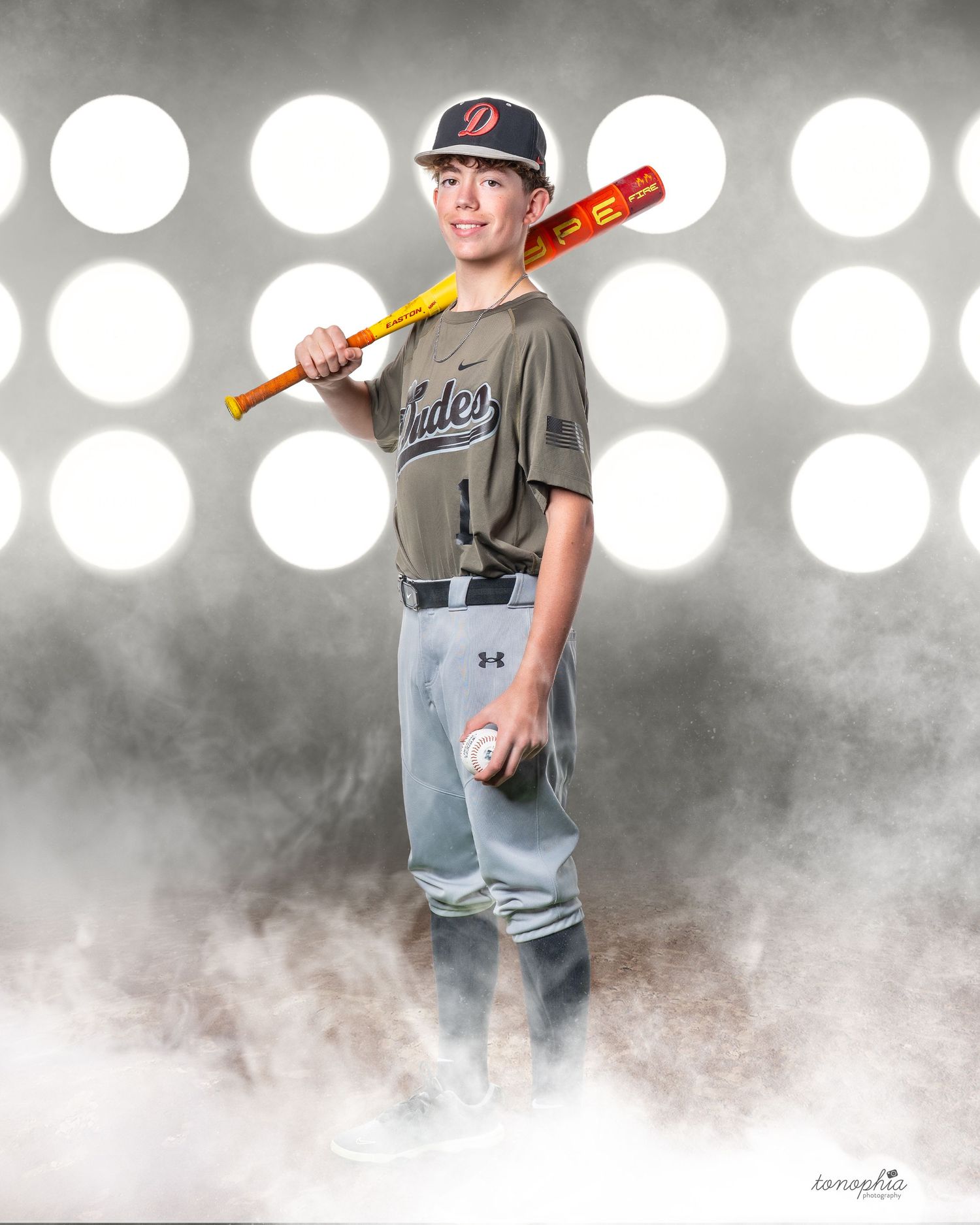 Youth baseball player in grey uniform demonstrates pitching stance against spotlit background.