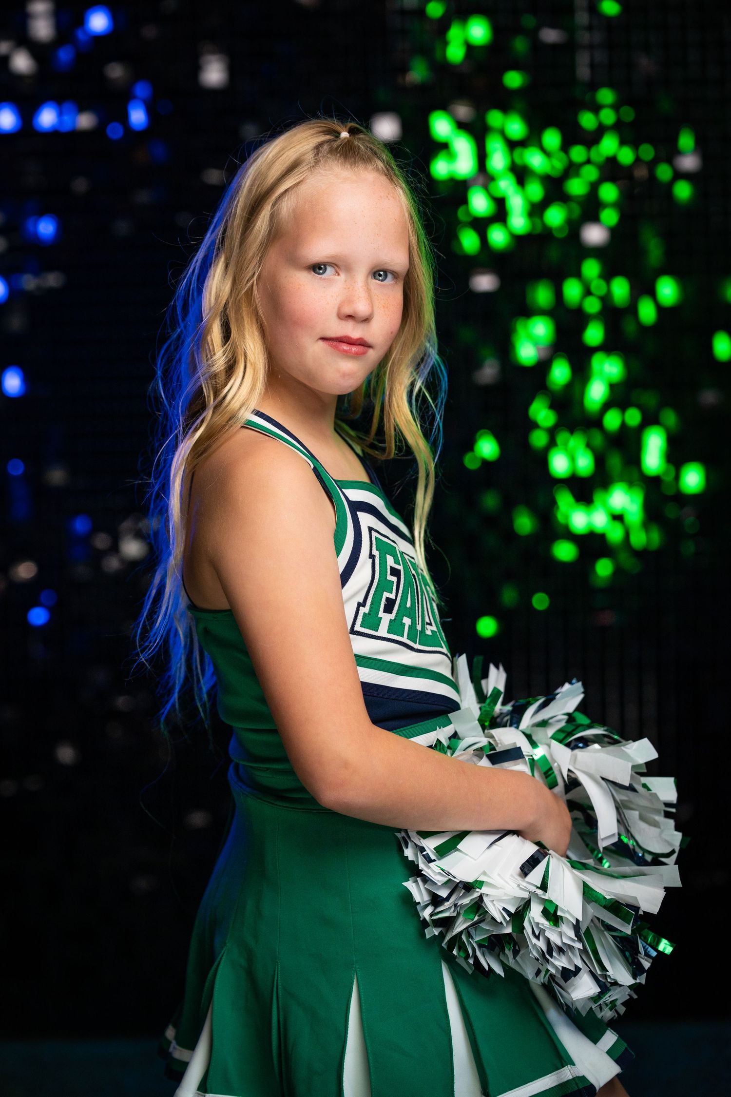 Cheerleader in green uniform with silver pom-poms performs against sparkly background.