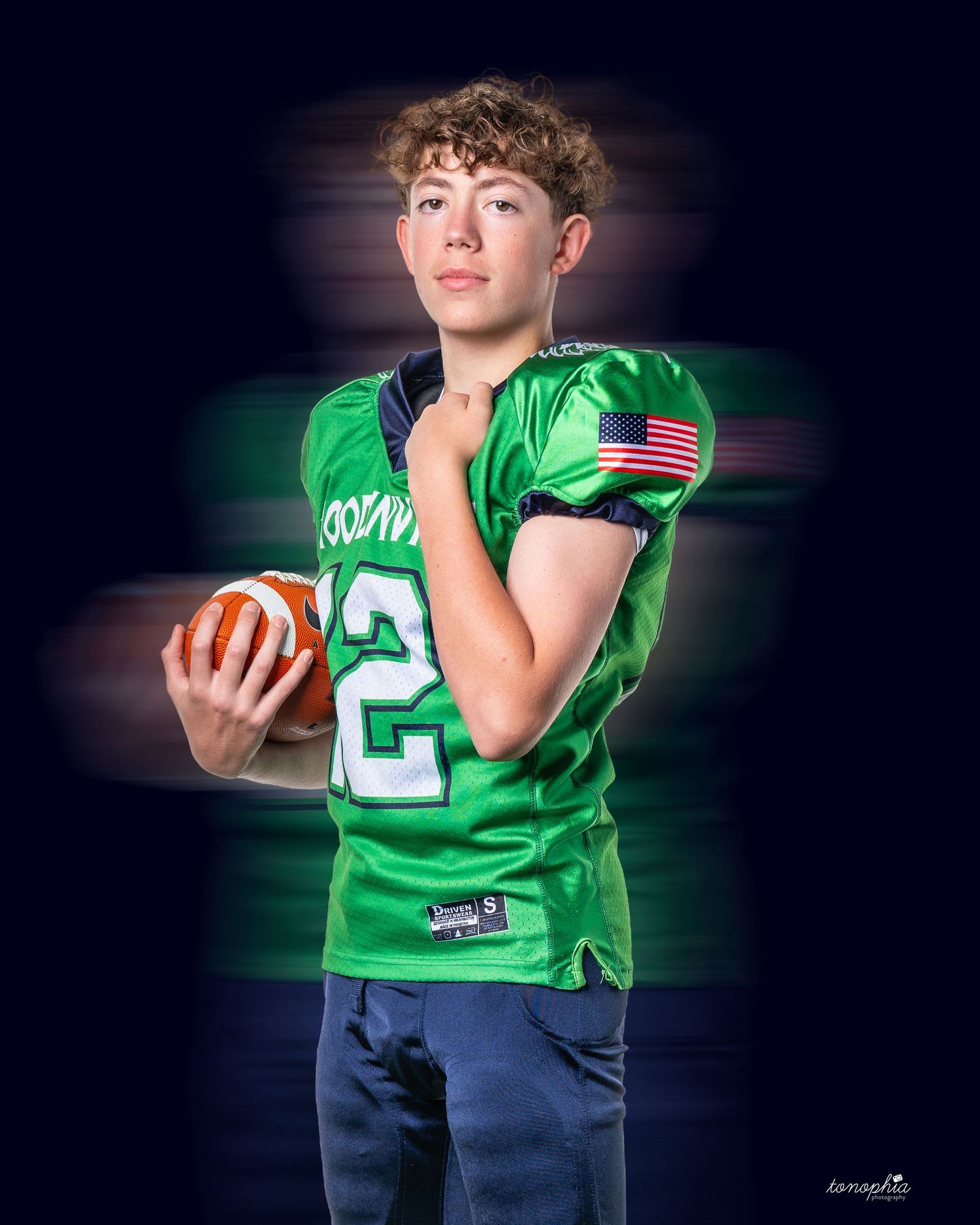 Youth football player poses with ball in green Woodinville uniform against dark background.