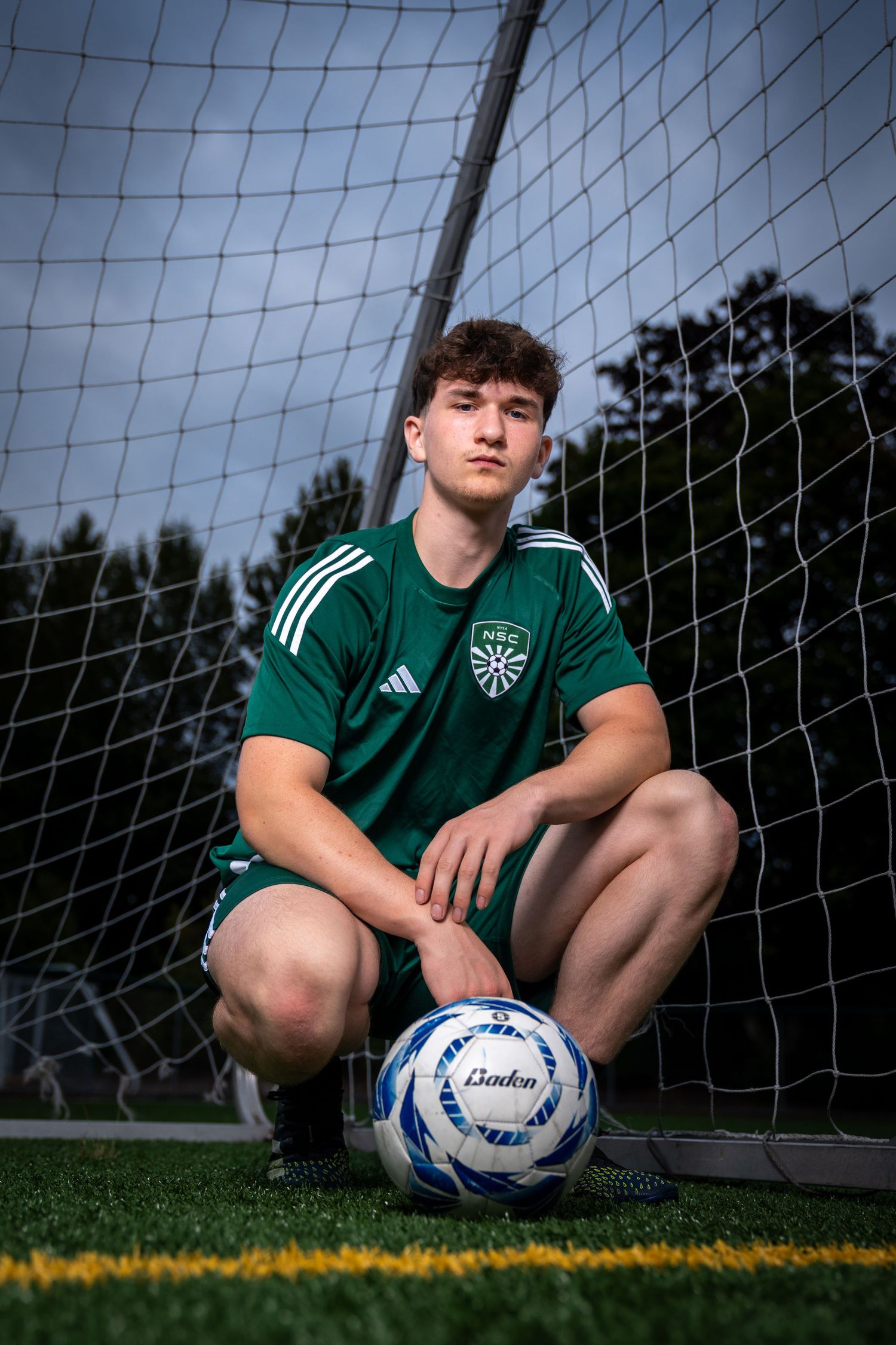 Soccer player in green jersey poses with ball by goal net on field.