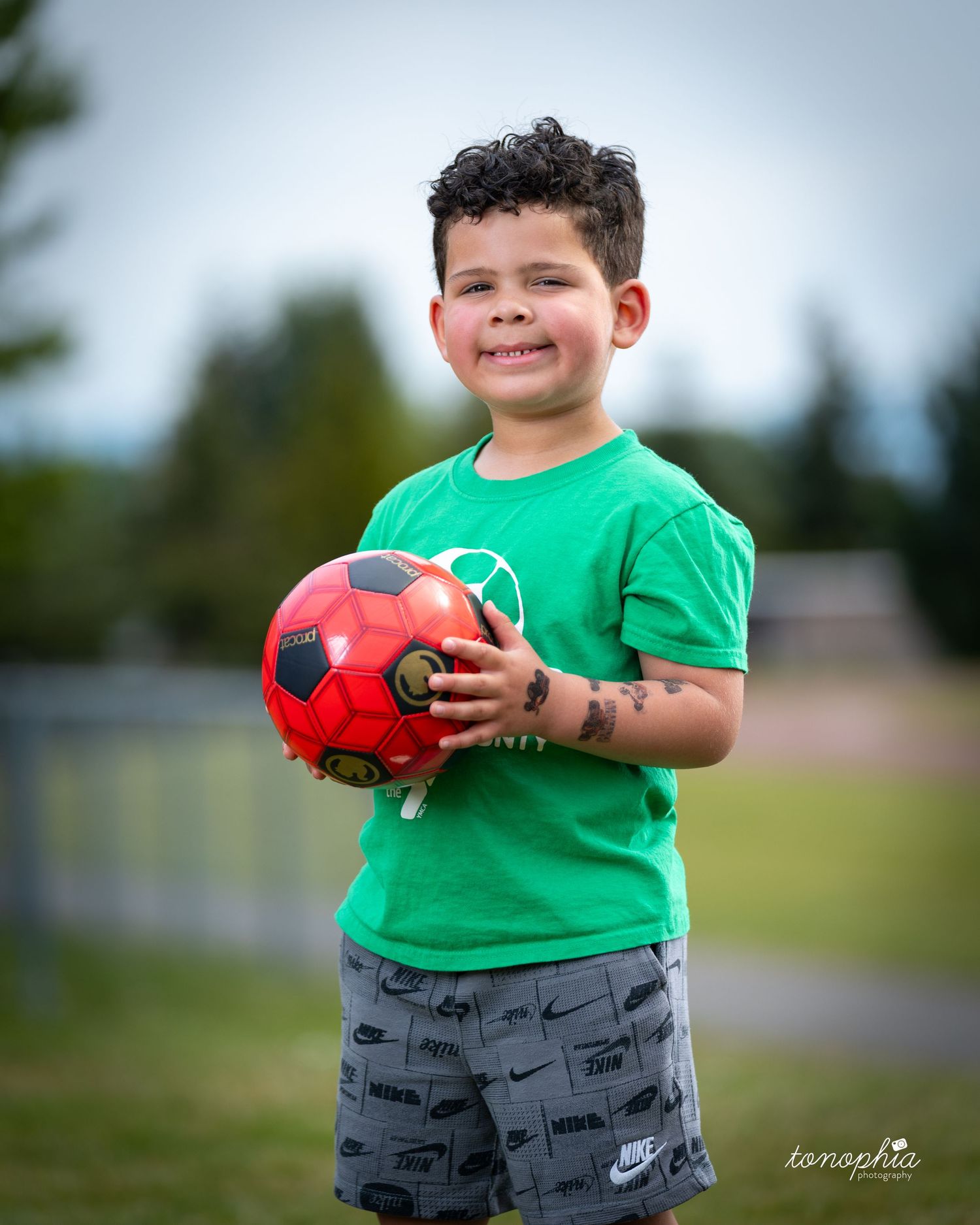 Young child in a bright green t-shirt holds a red and black soccer ball while standing outdoors on a sunny day.
