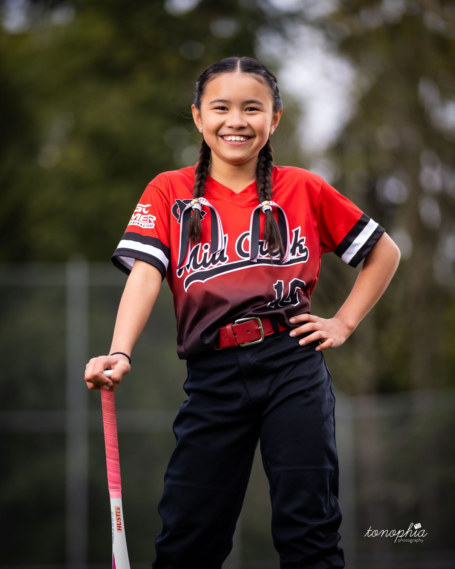 A softball player in a red and black uniform poses with a bat on a sunny day on the field.