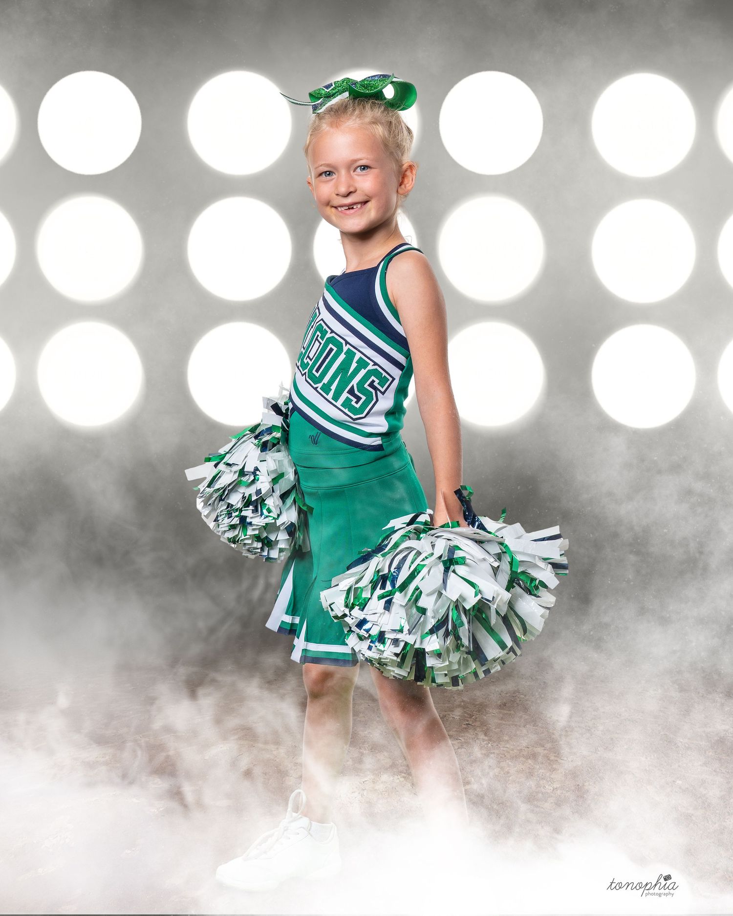 Youth cheerleader poses with green pom poms in front of spotlit background with fog effects.