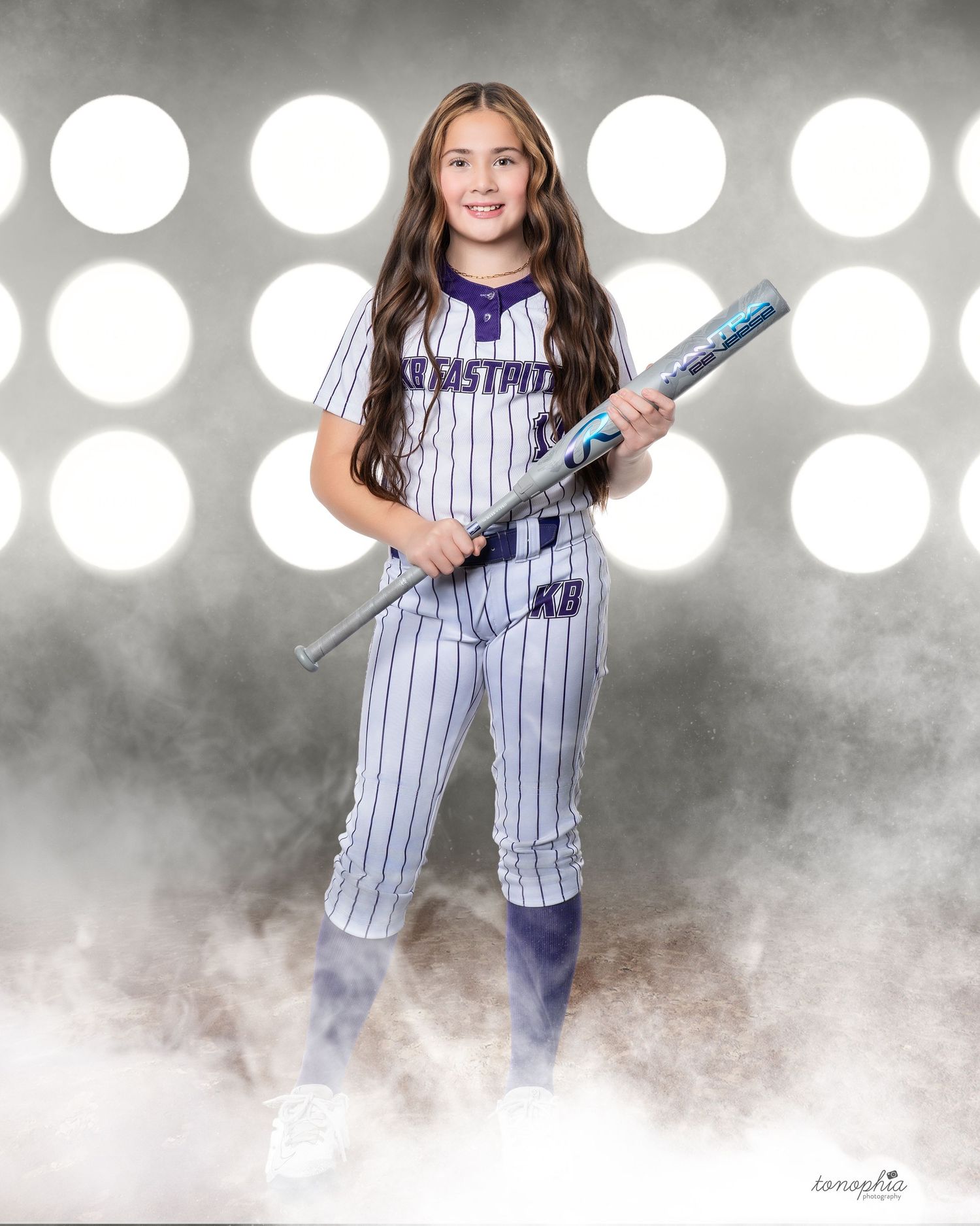 A young athlete in a pinstriped baseball uniform poses with a bat against a dramatic smoky background with spotlights.