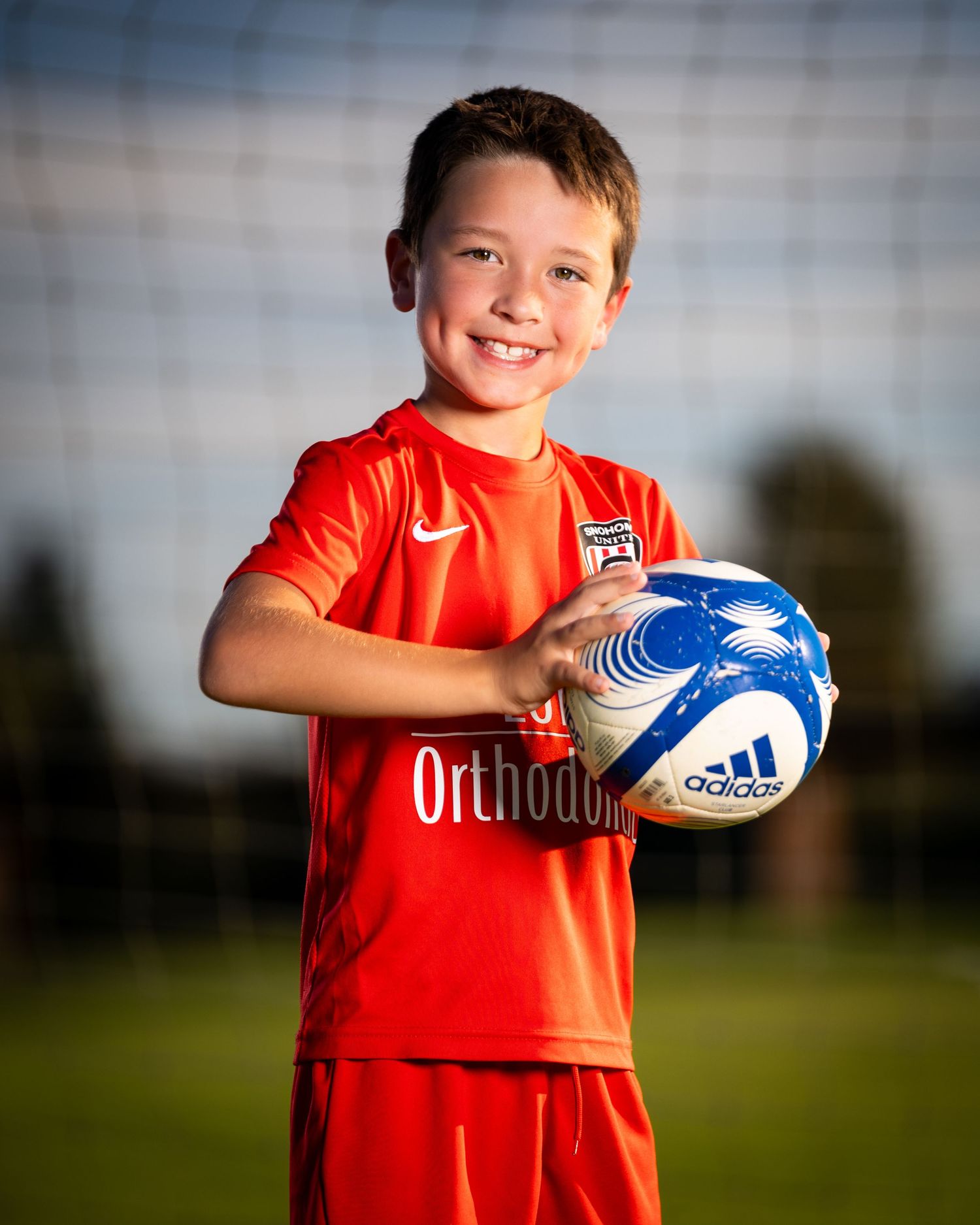 Young rugby player in a red jersey holds a blue and white rugby ball while smiling outdoors on a sunny day.