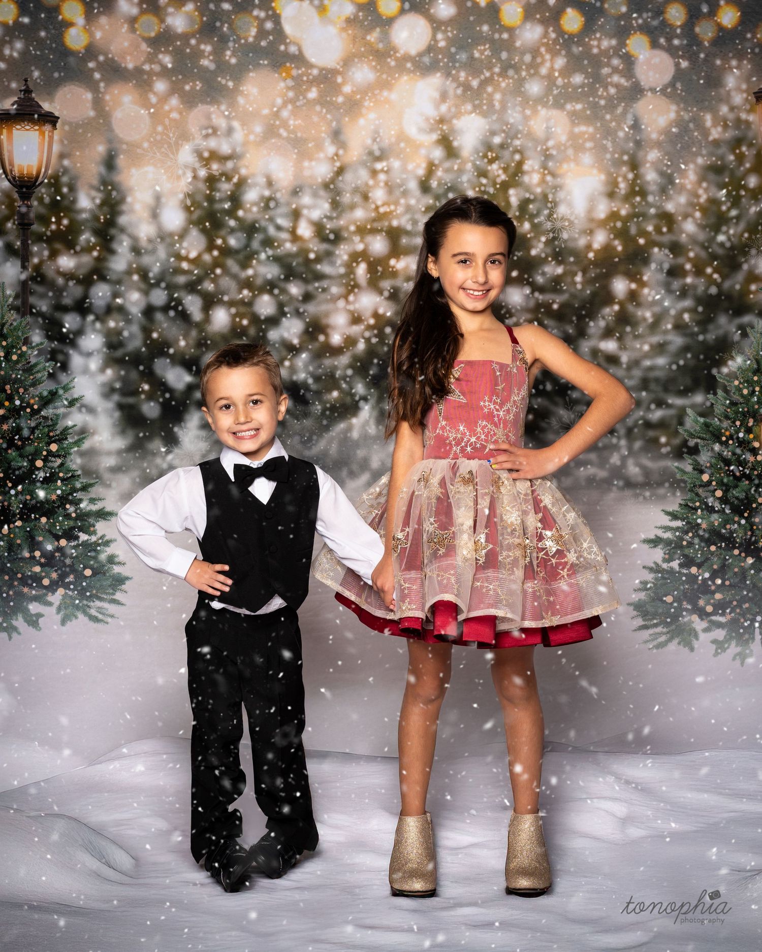 Festive winter photoshoot with children in formal attire against snowy backdrop with falling snow effect.