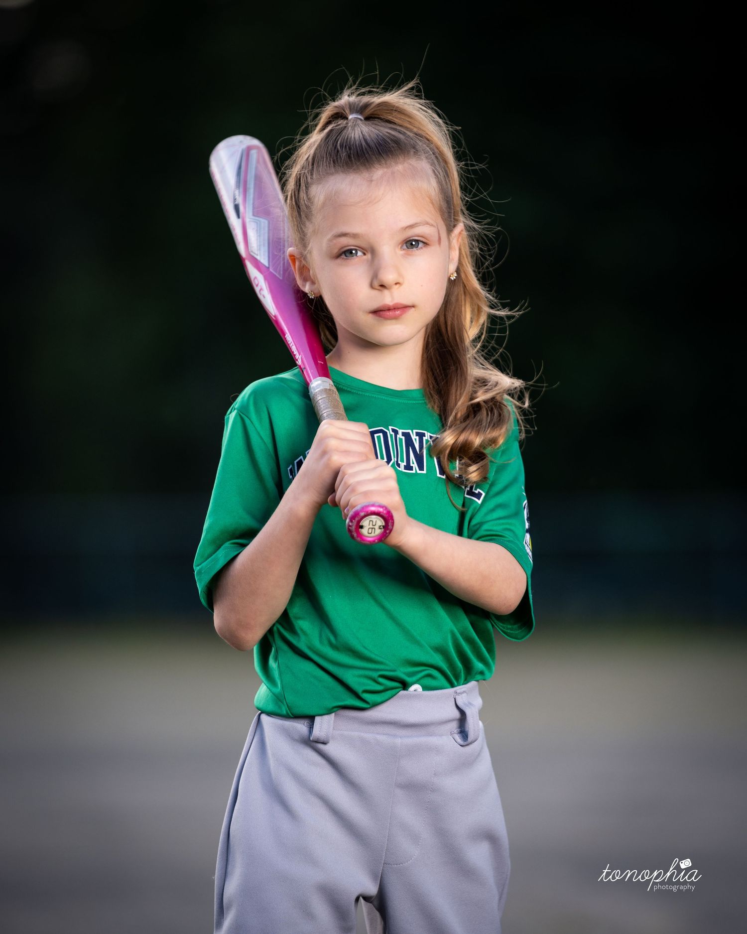 Young softball player in green jersey holds pink bat while posing for sports portrait outdoors.