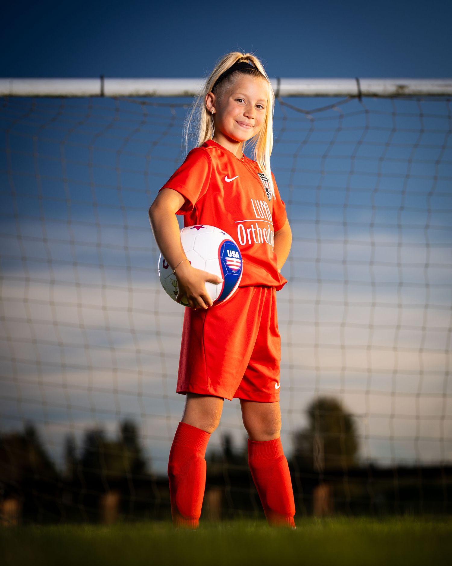 Soccer player in red uniform stands confidently against a goal net during an outdoor photo session.
