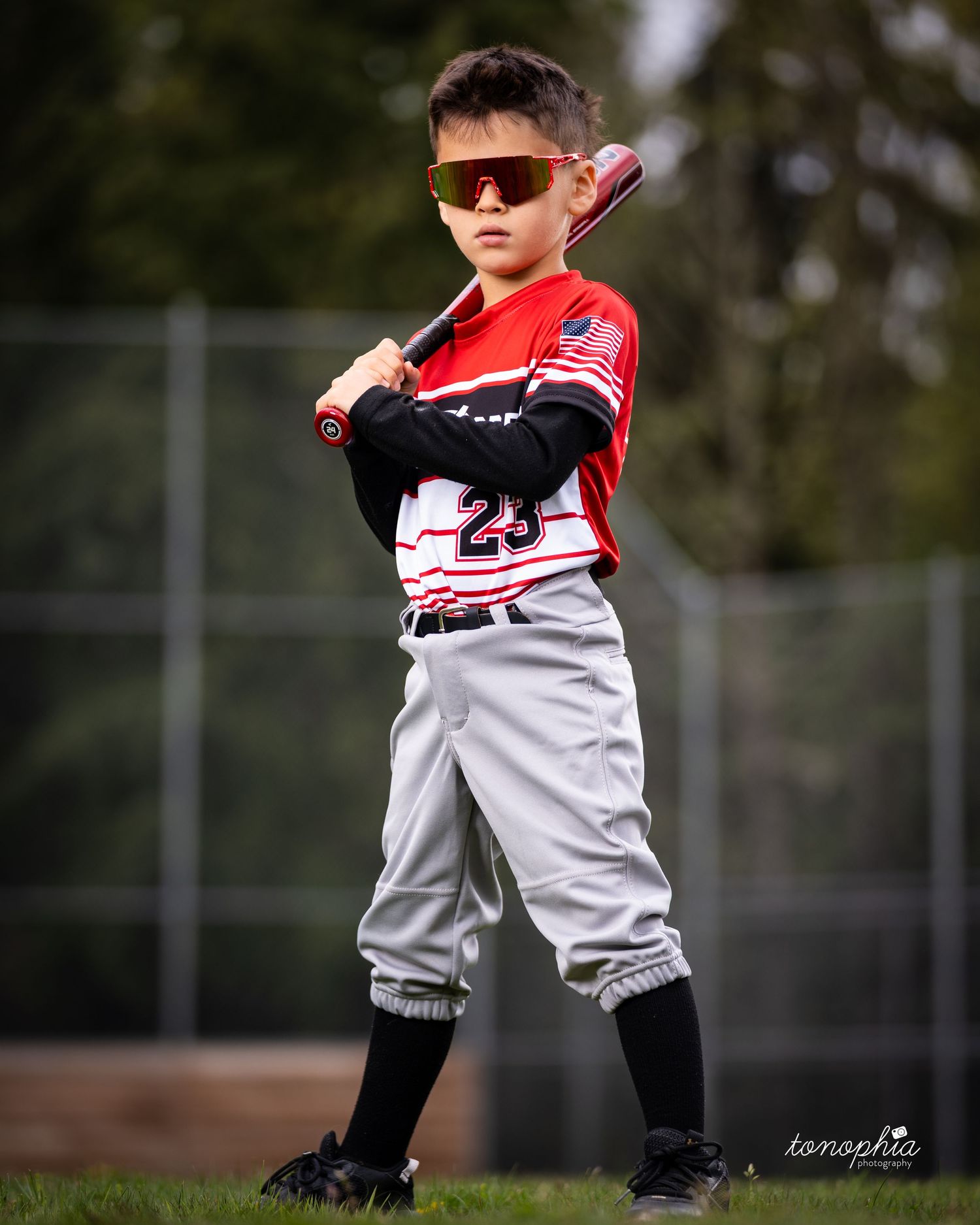 Young baseball player in red and white uniform strikes a dramatic pose during outdoor sports photography session.